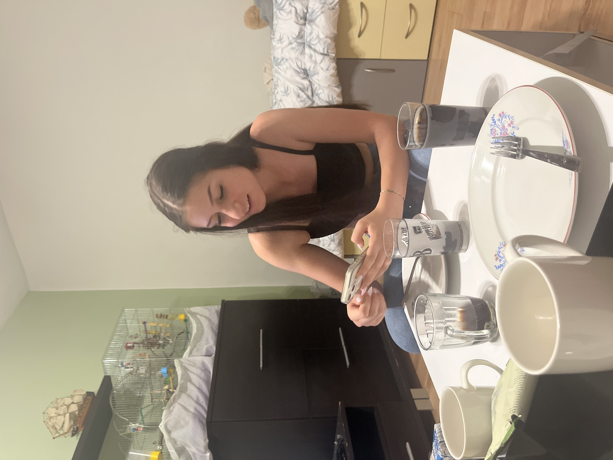 A young woman is seated at a white table, likely indoors, and is looking down at a smartphone in her hands. She is wearing a black top and jeans. The table is set with a plate, fork, a few glasses, and mugs. The background includes furniture like a wardrobe and a chest of drawers. A birdcage is also visible in the background. The city and specific location are unknown.