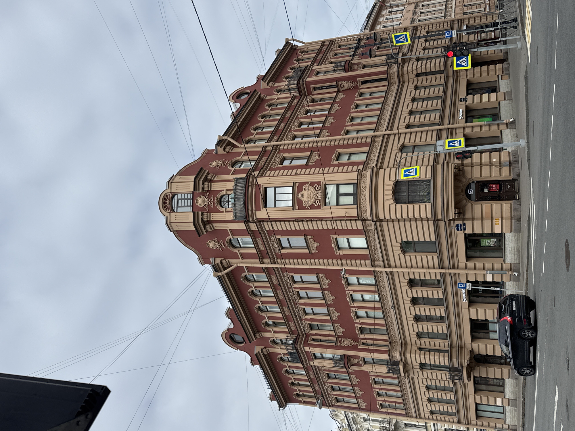 The image shows a corner building in Saint Petersburg, Russia. The building has a predominantly red facade with ornate beige trim around the windows and along the architectural features. The structure appears to be several stories tall with multiple rows of windows. A car is parked on the street adjacent to the building. There are pedestrian crossing signs and traffic lights visible. The sky is overcast.