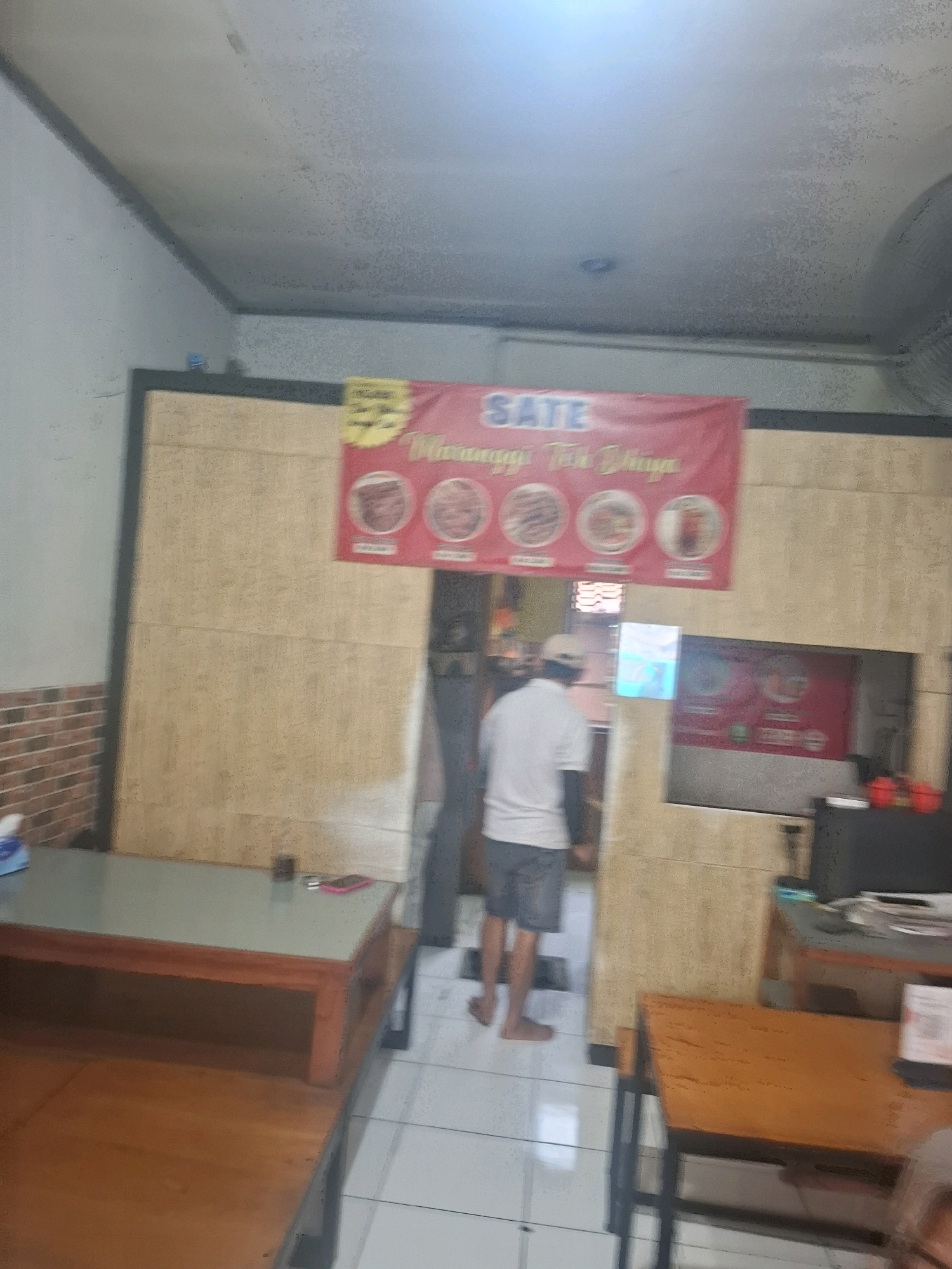The image depicts the interior of a casual eatery, likely a satay restaurant. A red banner with the word "SATE" prominently displayed in blue lettering hangs on a light brown wood-paneled wall. Below "SATE," smaller text in yellow reads "Maranggi Teluk Buyat." Several circular images of food are depicted on the banner.
A person wearing a white shirt, grey shorts, and a light-colored cap is walking away from the camera towards the right side of the frame. They are barefoot.
In the foreground on the left, a low wooden table with a glass top is visible. On the table, there are a few items including a tissue box, a dark object, and a pink smartphone. To the right of this table, another wooden table with a dark metal frame is partially visible.
Further into the establishment, a doorway leads to another area where a television screen displays a bright blue image. Another red banner, similar to the one at the top, is visible on the wall to the right of the doorway.
The environment appears to be dimly lit, with a textured ceiling and light-colored tiled flooring. The overall impression is a humble, possibly family-run, food establishment. The presence of satay-themed decor and the name "Maranggi" suggests a location in West Java, Indonesia, where Maranggi satay is a well-known dish. Given the context of the location being Cijalingan, Indonesia, this is likely a restaurant serving local Indonesian cuisine.
