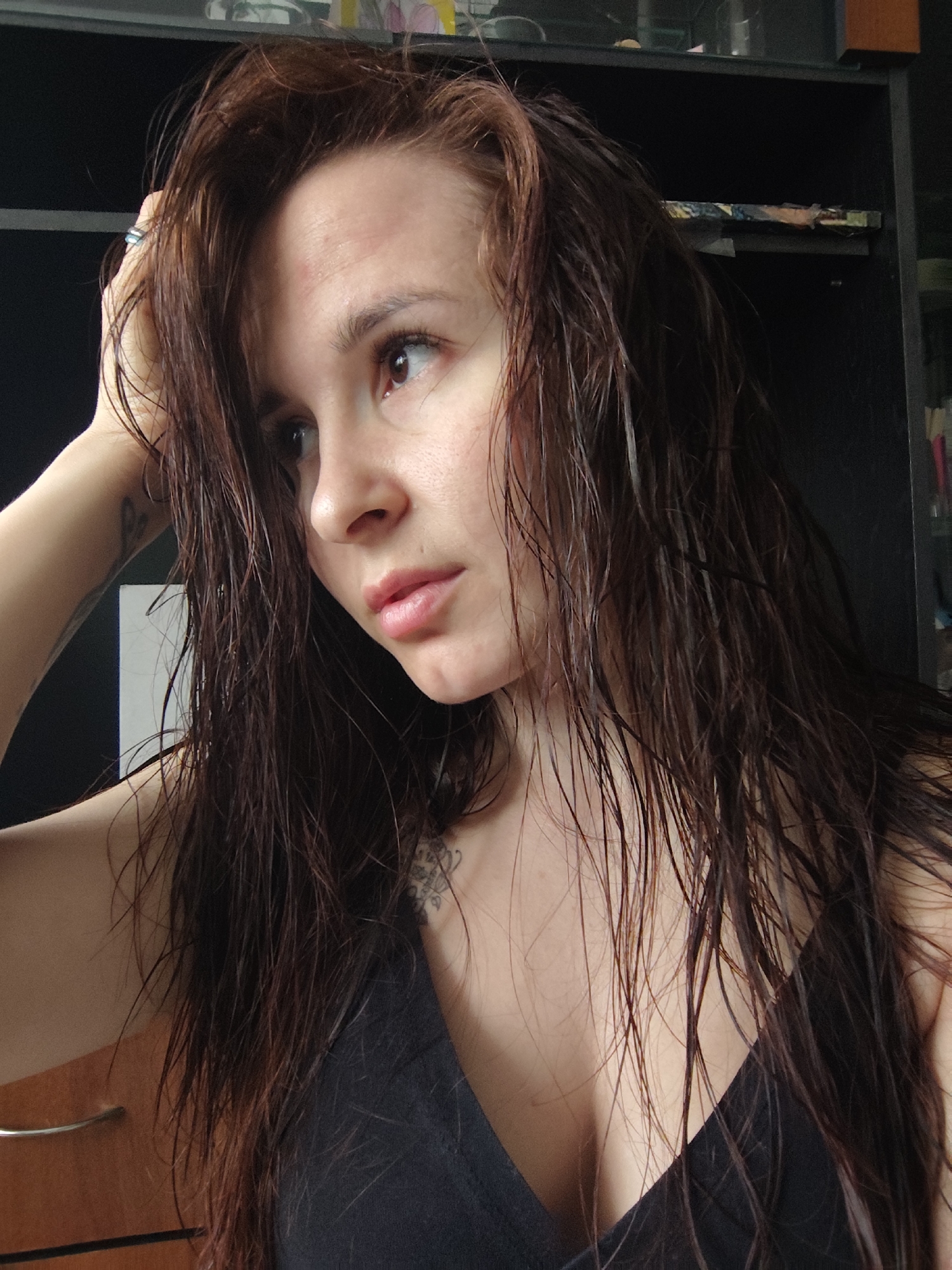 A medium close-up shot shows a woman with long, wet, auburn hair. She is fair-skinned, and her hair is slicked back on the top of her head. She is wearing a black tank top. Her left hand is raised towards her hair. A tattoo is visible on her left arm. Behind her is a dark, possibly black, cabinet. A wooden drawer is visible on the lower left. The location in the city of Фоки, Russia cannot be determined from the provided image.