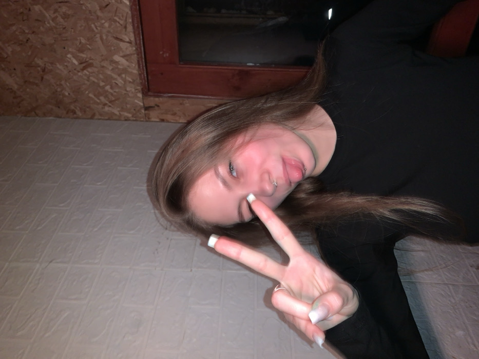 A young person with long, straight, light brown hair is the central focus of the image, which is rotated approximately 90 degrees clockwise. The person is lying down, looking up towards the camera with a playful expression. Their head is tilted slightly, revealing blue eyes, and they are sticking their tongue out. A silver-colored lip piercing (likely a vertical labret or similar) is visible on their lower lip, and a septum piercing (ring) can be seen in their nose. Their cheeks show a natural flush or blush.
With their right hand, they are making a "peace" sign (V-sign), holding it up towards their face. Their nails are long, shaped squarely, and feature a white tip (resembling a French manicure). They are wearing a plain black, long-sleeved top or shirt.
The background is divided into two main sections due to the image rotation. On what appears to be the left/top side of the image, there is a light-colored surface with a repetitive, textured pattern of small, rectangular raised sections, possibly a padded wall, floor tiling, or decorative panels. On the right/bottom side of the image, there is a wooden wall or paneling, possibly made of oriented strand board (OSB), with a visible reddish-brown wooden frame of what appears to be a window. Beyond this frame, the area is dark, suggesting it might be night outside or a dark interior.
The lighting is somewhat dim and artificial, casting soft shadows and highlights, particularly on the person's face and hair. The overall mood conveyed is casual, playful, and informal. No text is visible in the image.