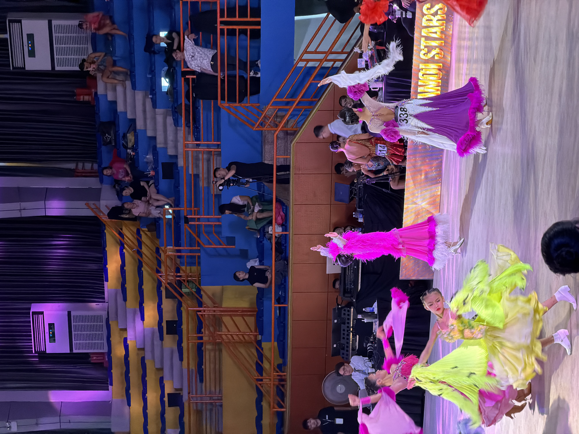 The image depicts a ballroom dancing competition at the Cung Thi Đấu Quần Vợt Mỹ Đình (My Dinh Indoor Games Arena) in Hanoi, Vietnam. Several young female dancers in brightly colored, elaborate gowns are performing on a polished wooden floor. The dresses are voluminous, with many layers and decorative elements like feathers and sequins. One dancer in a yellow and green dress is in the foreground, captured mid-spin with her skirt dramatically fanned out. Another dancer in a pink gown is to her left, also in motion. In the background, a stage is set up with lighting and what appears to be audio equipment. Spectators are seated in tiered blue and yellow bleachers, some observing the performance, while others are engaged with their phones. A large air conditioning unit is visible on a raised platform behind the stage. The overall atmosphere suggests a formal competition or performance event.