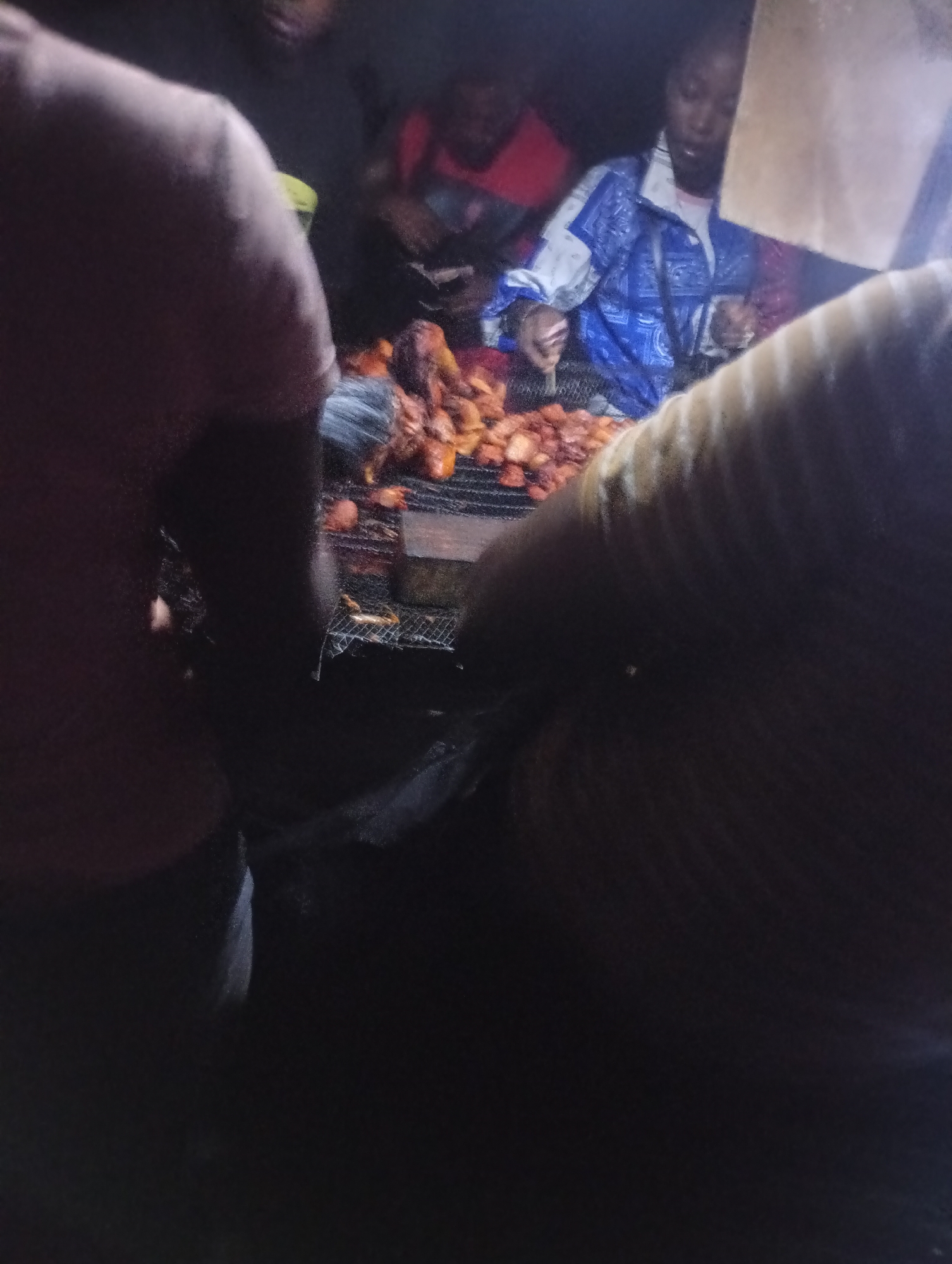 In Nkpor, Nigeria, a group of people are gathered around a charcoal grill at night. Several individuals, including a woman in a blue and white patterned jacket and a person in a red shirt, appear to be preparing or serving food. The food, which resembles fried chicken or plantains, is laid out on a metal grate over the coals. The scene is dimly lit, with a focus on the cooking area. The foreground is dominated by a large, out-of-focus object, possibly a basket or woven mat, casting a shadow over the scene. The overall impression is that of a busy, nighttime food stall or outdoor cooking event.