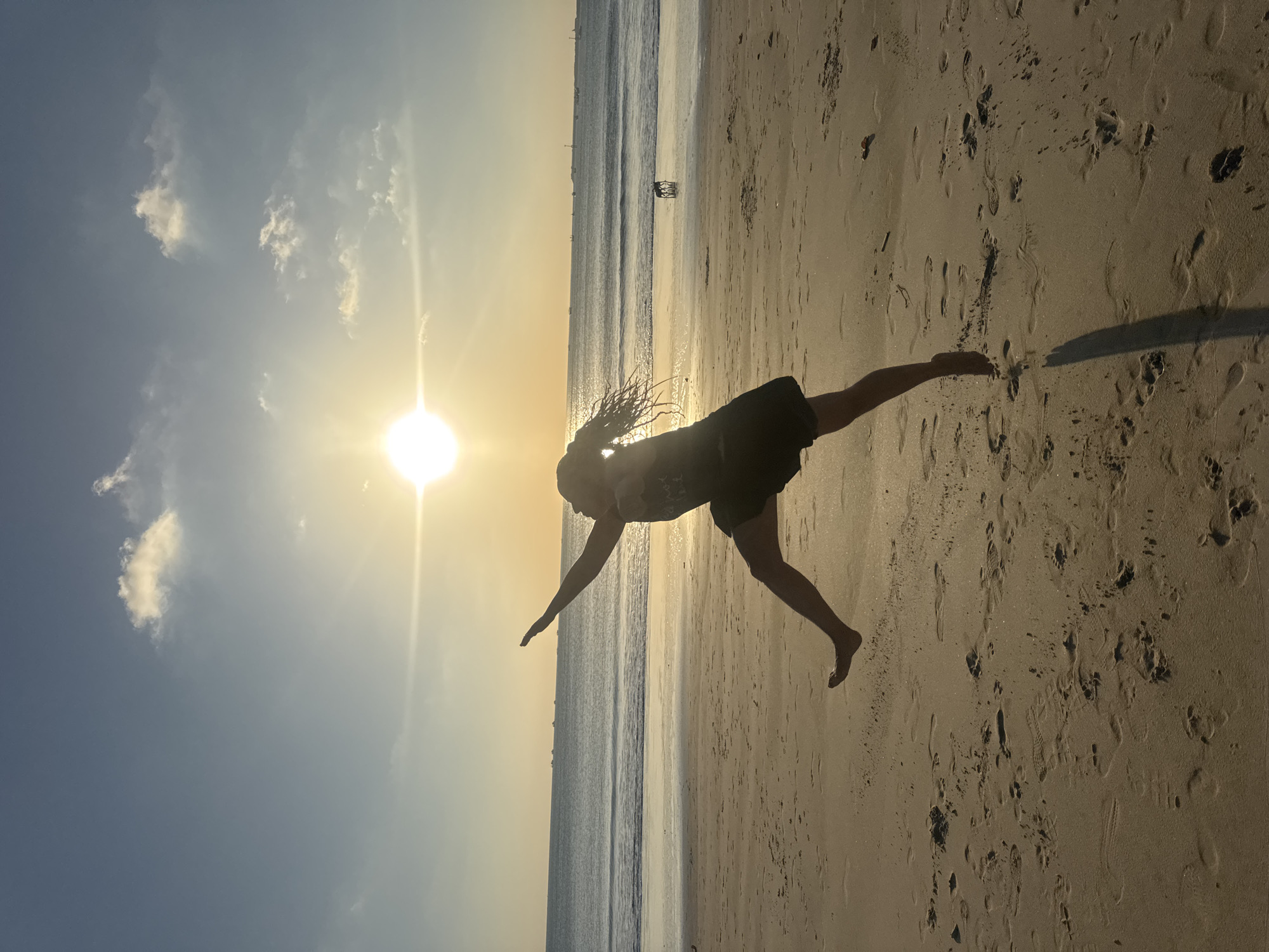A person is captured mid-jump on a sandy beach in Kuta, Indonesia, during sunset. The person, silhouetted against the bright sun, has their arms outstretched and legs in a dynamic pose, suggesting joyful movement. The sun, low on the horizon, casts a warm glow over the scene, illuminating the gentle waves of the ocean and the vast expanse of the sky. The beach is sandy, with footprints indicating recent activity. In the distance, several boats are visible on the water, and two dogs can be seen near the shoreline. The overall atmosphere is serene and vibrant, embodying the beauty of a tropical sunset.