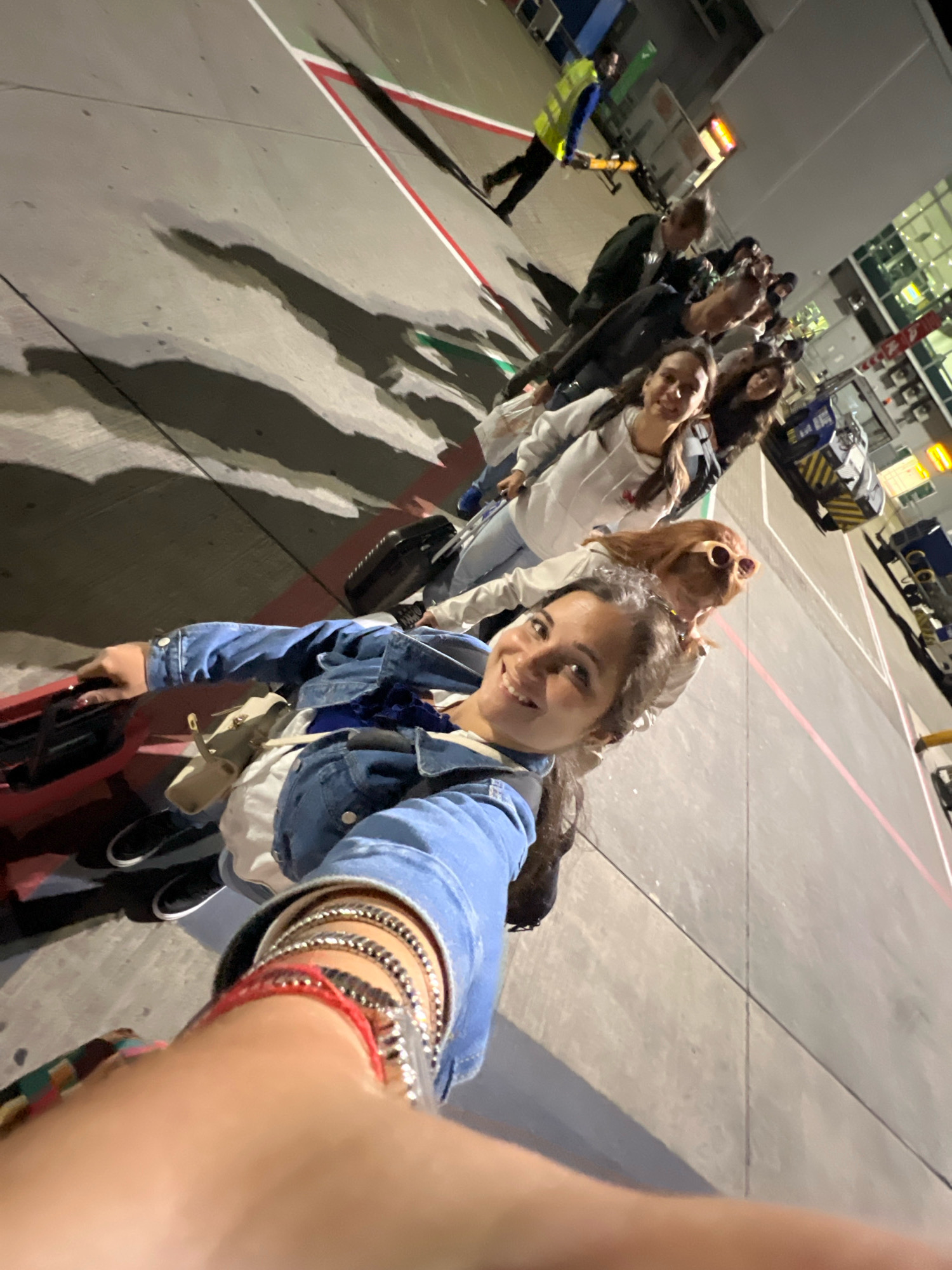A group of people are walking in a line on an airport tarmac at night, seemingly boarding an airplane. A selfie is being taken by a woman in the foreground, who is wearing a denim jacket and bracelets. She is smiling at the camera. Behind her, other individuals, including women in casual attire, are visible, some pulling luggage. A person in a high-visibility vest is further down the line, suggesting they are airport personnel. The tarmac is marked with white and red lines. The background shows parts of airport buildings and lighting. The overall scene appears to be an organized boarding process at an airport in Uttlesford, United Kingdom.