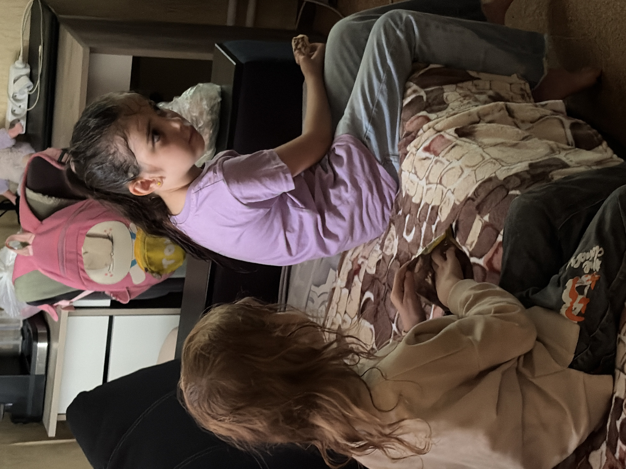 Two young girls are seated on a bed, covered by a patterned blanket, in what appears to be a domestic setting. The girl on the right, facing the viewer, has dark hair pulled back in a ponytail and is wearing a light purple t-shirt and blue jeans. She is looking to her left and holding a piece of food in her right hand. The girl on the left, with light brown wavy hair, is facing away from the viewer and is wearing a light tan sweatshirt and dark pants with an orange and white graphic on the left leg. She is looking down at her hands, which are resting on the blanket, and appears to be holding a snack or a wrapper. In the background, a pink backpack with a cartoon face is visible, along with furniture that suggests a room in a home. The scene is not demonstrably located in Tambov, Russia, as there are no specific indicators to confirm this location.