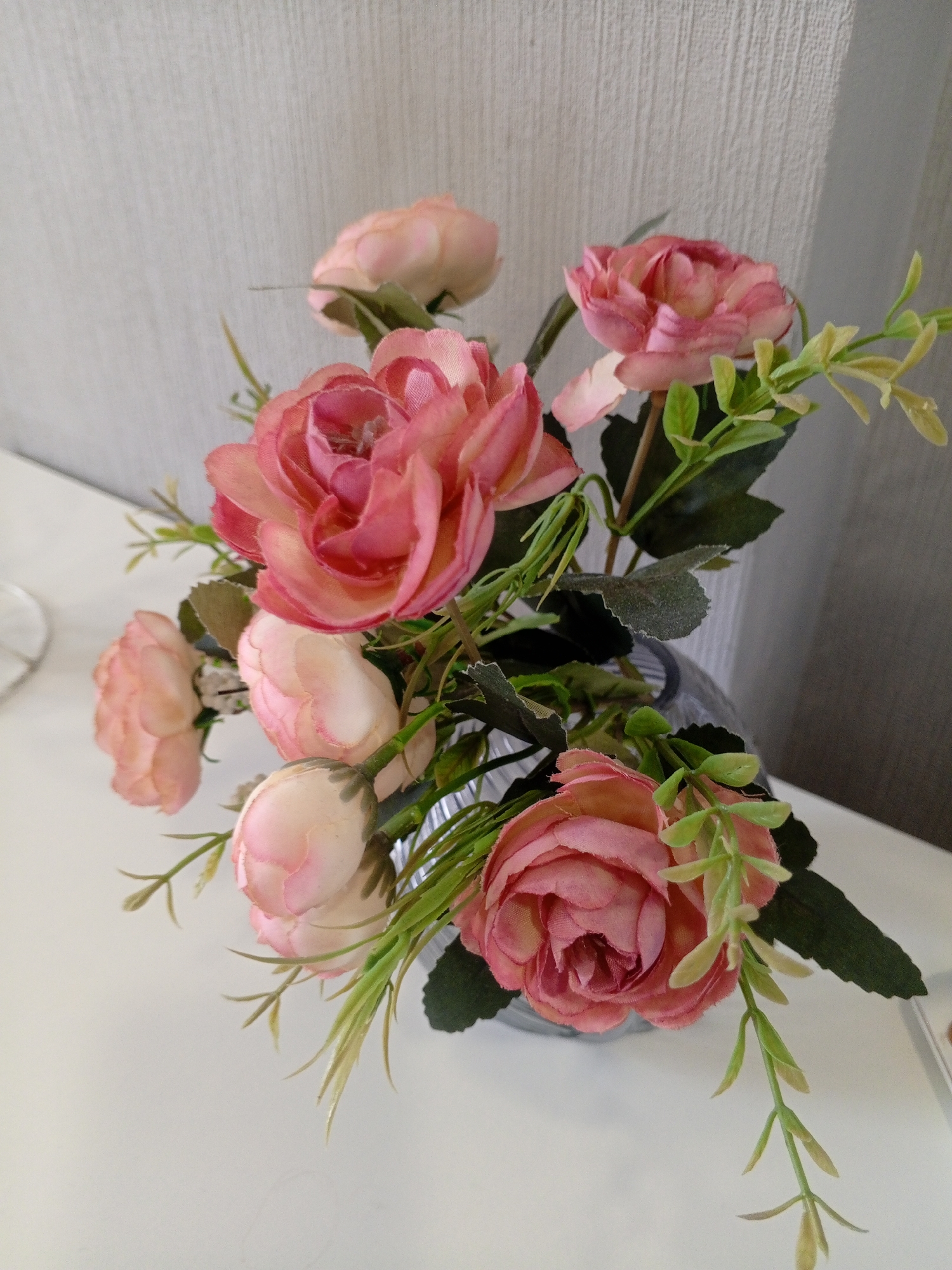 A bouquet of artificial pink roses and greenery is arranged in a clear glass vase. The bouquet is placed on a white surface, likely a table or shelf. In the background, a textured, light-colored wall can be seen. The lighting appears to be natural, casting soft shadows. There are no people or animals present in the image. The setting does not provide any specific details that would place it in the city of Sochi, Russia.