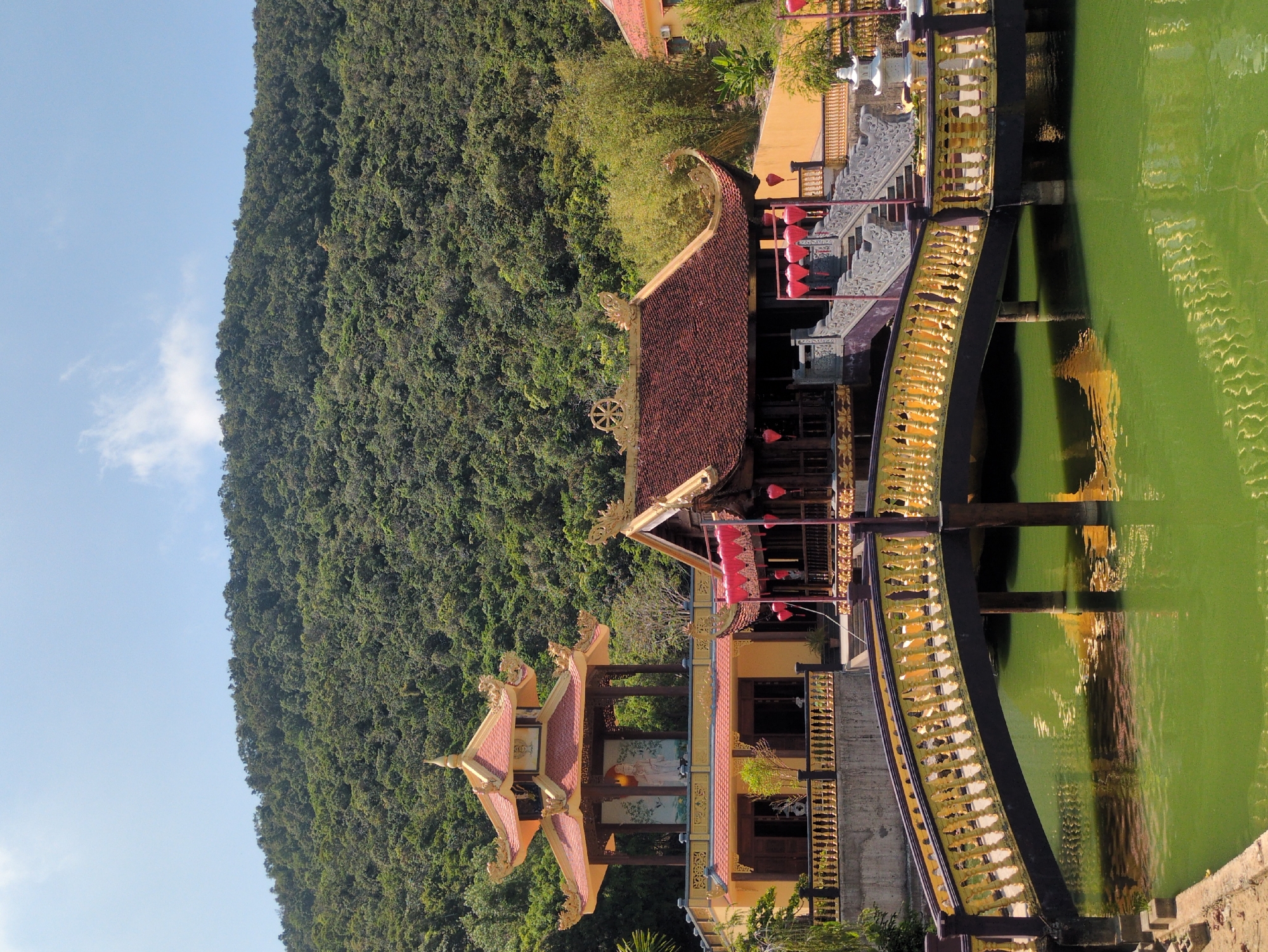 View of Hùng Long Pagoda, Phú Quốc, Vietnam. A hillside covered in trees is to the left. To the right are buildings and a golden arched bridge over a green pond. There is a clear sky.