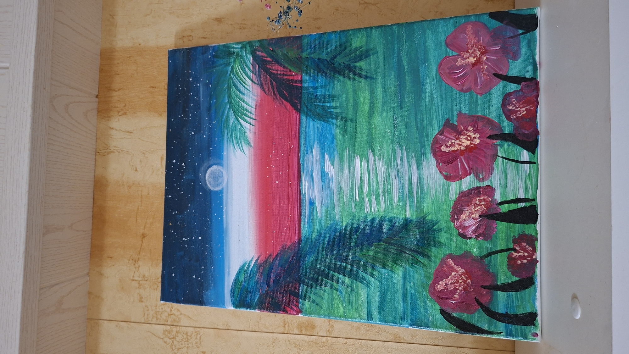 The image displays an acrylic painting on canvas. The painting depicts a tropical night scene with a starry sky, a moon, palm trees, and a body of water. In the foreground, there are several large pink flowers with dark stems. The painting is positioned on a white shelf against a textured yellow wall. There is no visible indication of the location being in the city of Большой Букор, Russia.