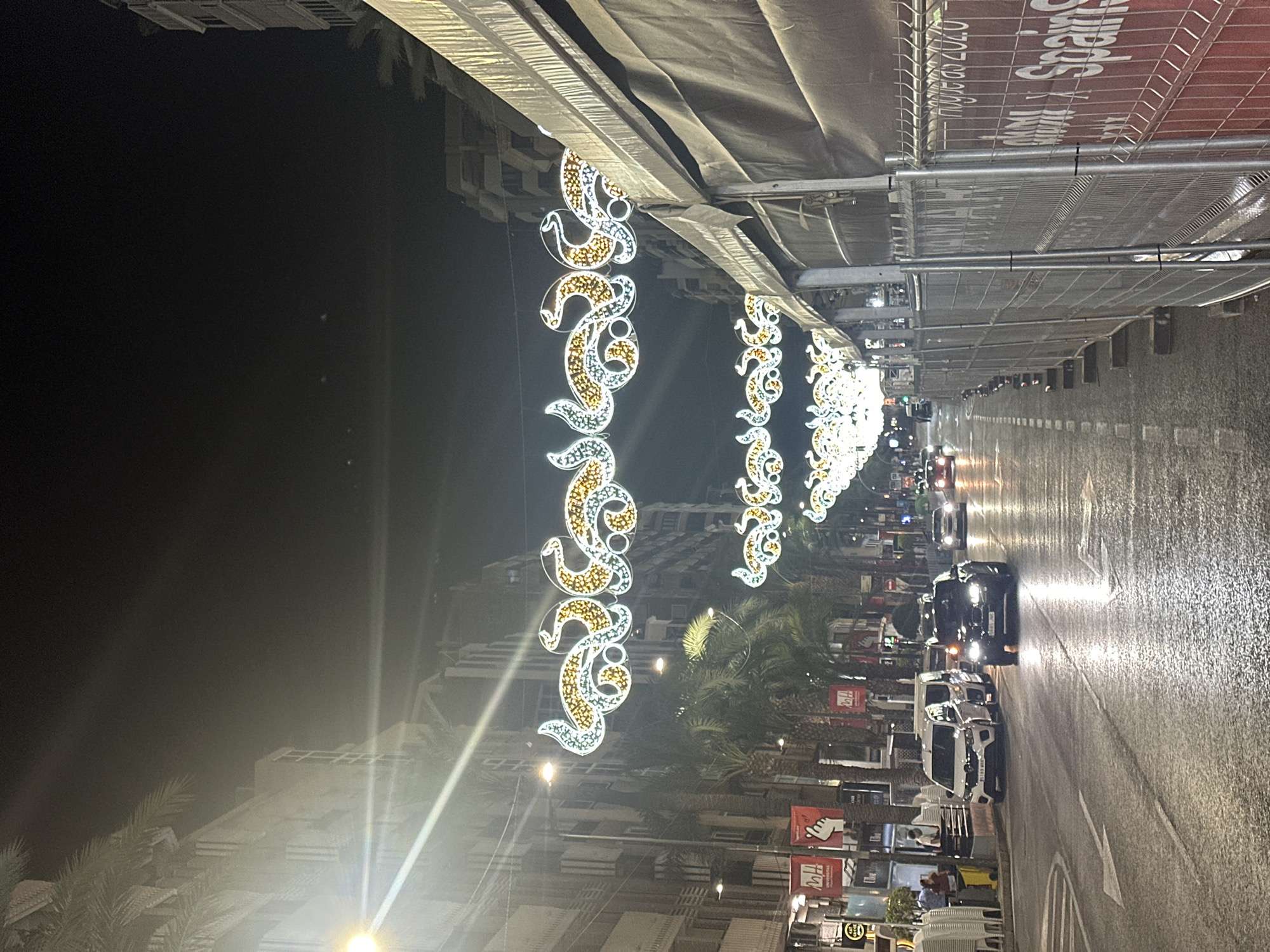 This image shows a nighttime street scene in Alicante, Spain, specifically on Rambla de Méndez Núñez, a prominent street known for its shops and festive decorations. The street is lined with multi-story buildings, palm trees, and streetlights. Overhead, decorative lights in the shape of sinuous, ornate patterns are strung across the street, illuminated by white and yellow lights, likely part of a seasonal celebration or event.
The street itself is wet, indicating recent rain or street cleaning, and reflects the lights from the streetlamps and vehicles. Several cars are visible on the road, some appearing to be parked and others in motion, their headlights cutting through the darkness. On the left side of the street, the facade of a building shows windows and architectural details. Various signs and banners are attached to lampposts and buildings, some displaying text and graphics. One visible banner on the left has the text "CAT CASA" and another red banner has a hand gesture symbol with text.
On the right side of the street, a temporary construction barrier, made of metal fencing and opaque sheeting, runs along the sidewalk. A large red banner is attached to this barrier, with text that appears to include "Spain" and "2020". A few people are visible on the sidewalk on the left, one standing near a yellow bin and another near a parked white van. The overall atmosphere is that of a lively urban street at night, enhanced by festive illuminations.