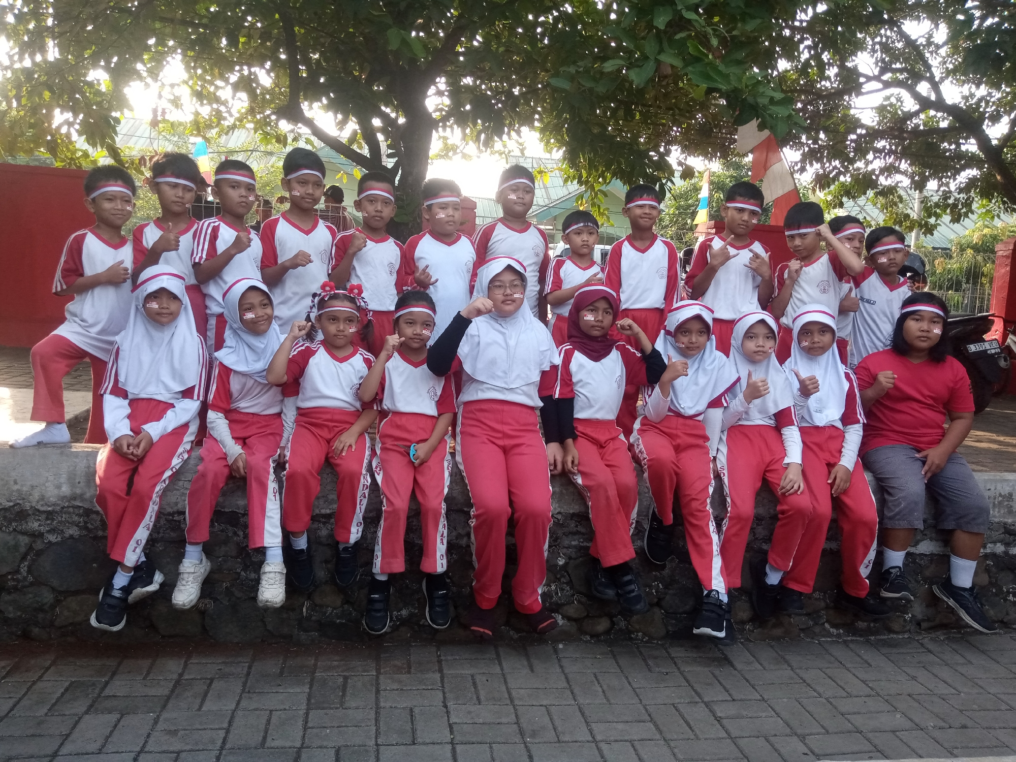 A group of Indonesian schoolchildren, both boys and girls, are posing for a photo outdoors. They are all wearing matching red and white uniforms, with white headbands adorned with red and white stripes. Many of them have red and white face paint, resembling the colors of the Indonesian flag. The children are arranged in two rows, with some standing and others sitting on a stone wall. They appear to be participating in a school event or celebration. The background shows trees with dappled sunlight filtering through, and a hint of a red and white flag on a pole, suggesting a patriotic occasion. The ground is paved with grey interlocking bricks. The scene takes place in Sukaraja, Indonesia.