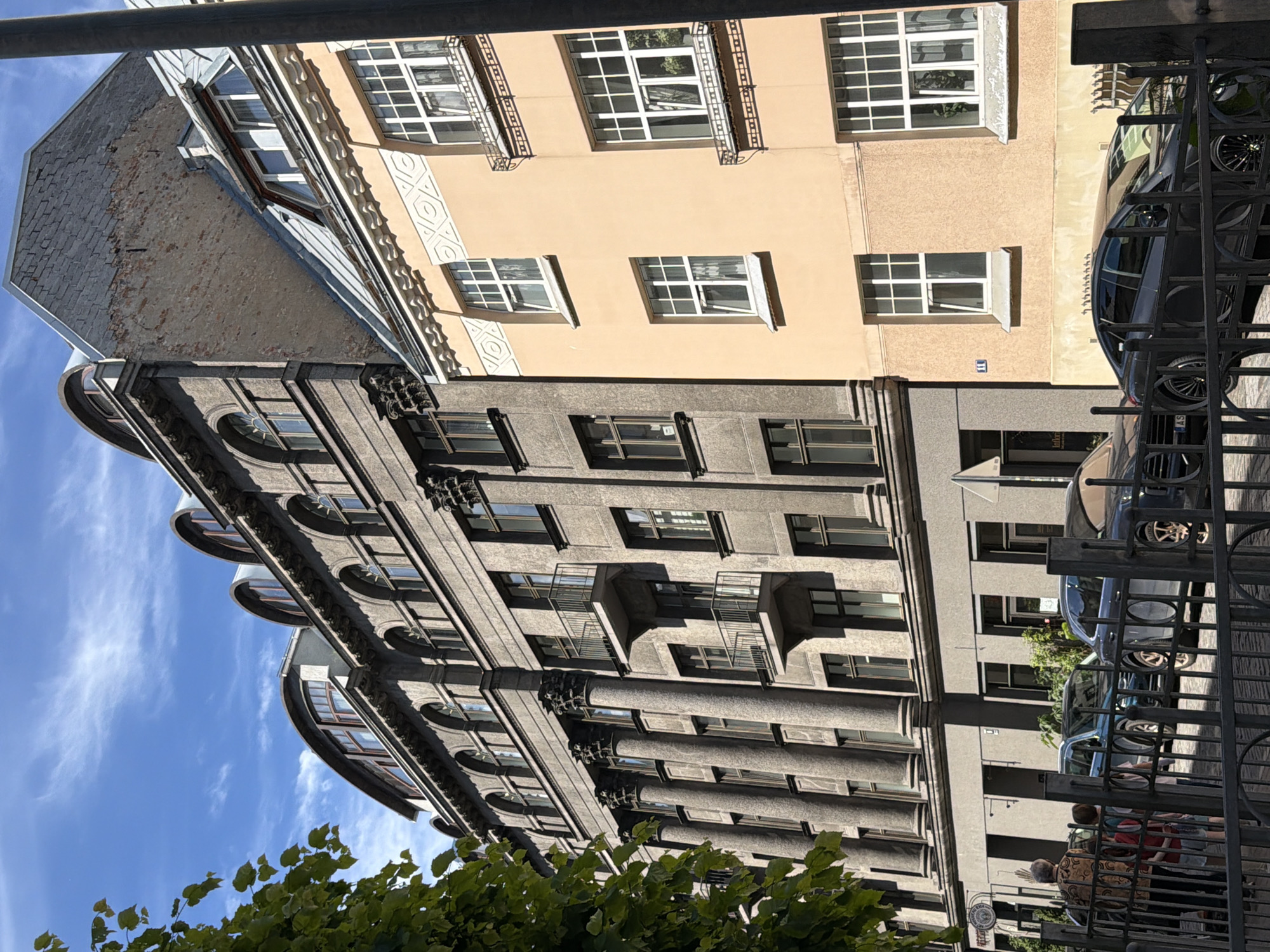 A sunlit, upward-angled exterior shot shows the facade of two adjacent buildings in Riga, Latvia. The building on the left is a dark gray, imposing structure with Corinthian columns. The building on the right is a lighter peach color with white decorative trim around its windows. The sky above is a bright blue with thin, wispy white clouds. In the foreground, a black metal fence separates the viewer from the street. Behind the fence, a dark gray sedan and a dark blue SUV are parked. To the left of the cars, three people are standing: an older individual wearing a patterned jacket and two children. The older individual is facing away from the camera, looking towards the buildings. The children are also facing away, looking in the same direction. Trees with green leaves are visible on the far left of the image.