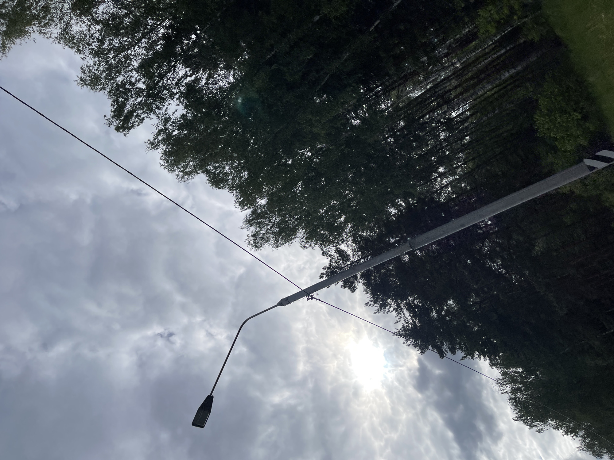 The image shows a street scene in Semyonovskoye, Russia, with a cloudy sky and sunlight breaking through. A street lamp extends from a utility pole, with power lines visible. Trees line the road, obscuring the view. A road sign is partially visible.