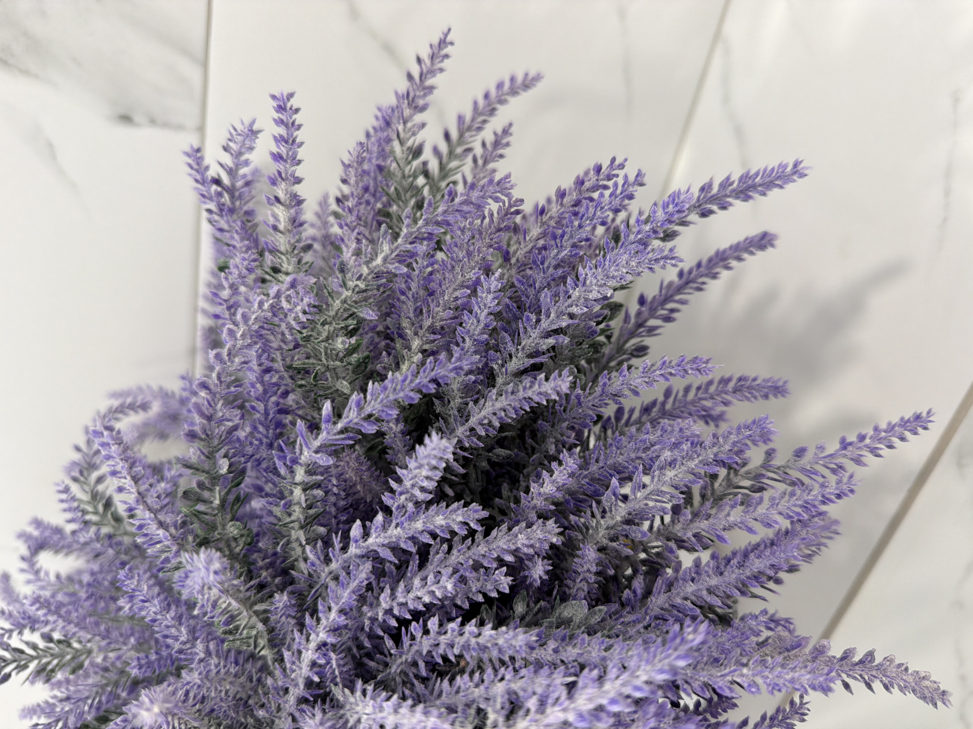 A close-up shot focuses on a bunch of artificial lavender flowers. The lavender is a vibrant purple color and has a slightly dusty or frosted appearance, suggesting it might be artificial. The flowers are arranged in a dense cluster, with some stems drooping downwards. The background consists of a white, marbled surface, possibly a countertop or a wall, with subtle grey veining. The lighting casts soft shadows from the lavender onto the background, indicating a light source from above and slightly to the left. The image is captured from a high-angle perspective, looking down into the arrangement of flowers. There are no people or any discernible context related to the city of Kazan, Russia, in the image.