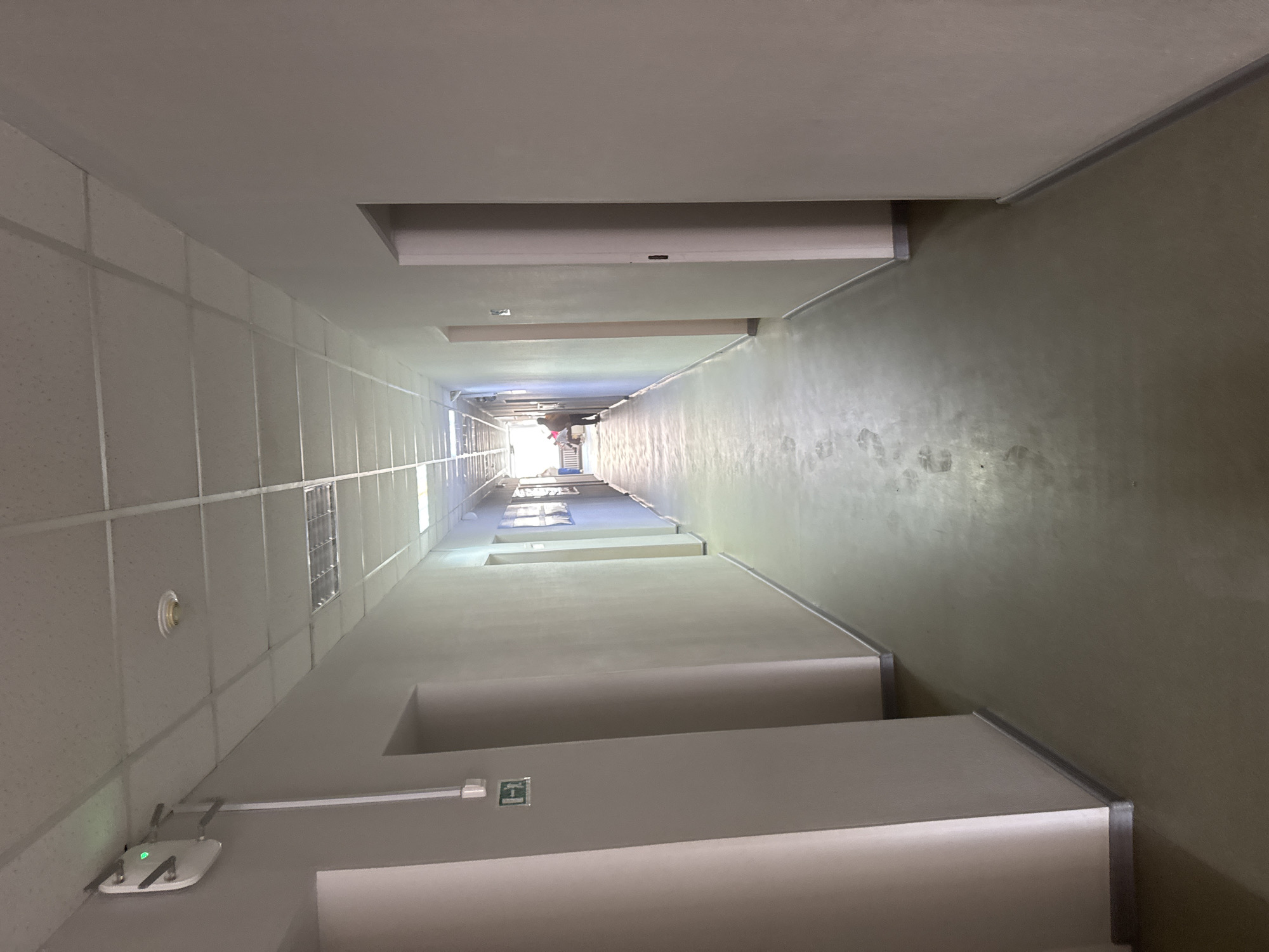 The media file captures a long, narrow stairwell or hallway, viewed from a low-angle looking straight up. The perspective emphasizes the height and verticality of the structure, creating a sense of a deep shaft.
On the left side, the wall features a light-colored grid-like pattern, possibly composed of acoustic panels or tiles. A round white object, likely a smoke detector, and a rectangular grid-patterned window or vent are visible on this wall. Further down, an access point or router with two antennae and a glowing green light is mounted near the base of the left wall.
The right wall is a smooth, light olive-green or beige color, showing what appear to be smudges or footprints ascending along its surface, suggesting it might be the underside of a staircase or a sloped wall that people might walk on. The stairs themselves are light-colored with prominent metallic edge strips, forming a multi-level structure that gradually narrows towards the top.
At the distant end of the ascent, a bright, intense light source is visible, suggesting an exit or an external, brightly lit area. A single person is faintly discernible near this light source, appearing to be standing or walking.
Overhead, multiple levels of ceilings are visible, with long fluorescent light fixtures providing illumination. A green square sign on the lower part of the left structure reads "Bblxog" (Russian for "Exit") with an arrow pointing right, confirming the location in a Russian-speaking region, specifically Perm, Russia. The image is devoid of specific activity beyond the presence of the distant person, focusing instead on the architectural features and perspective.