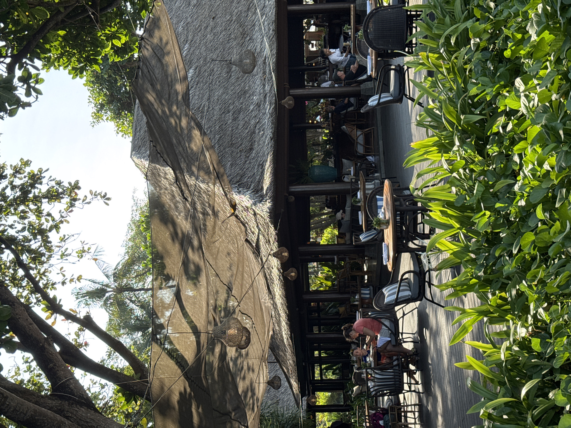 The image depicts an outdoor seating area of a restaurant or cafe, possibly in Denpasar, Indonesia, under a thatched roof structure with a large, draped fabric overhead providing shade. Lush green foliage is visible in the foreground, with bright sunlight filtering through trees onto a wooden deck. Several tables with chairs are set up, with some patrons seated and dining. In the middle ground, two individuals are seated at a table, with one appearing to be a child and the other an adult wearing an orange shirt and white shorts, looking down at something on the table. In the background, other patrons are visible, and a server in a white shirt is attending to a table. The overall atmosphere suggests a relaxed dining experience in a tropical setting.