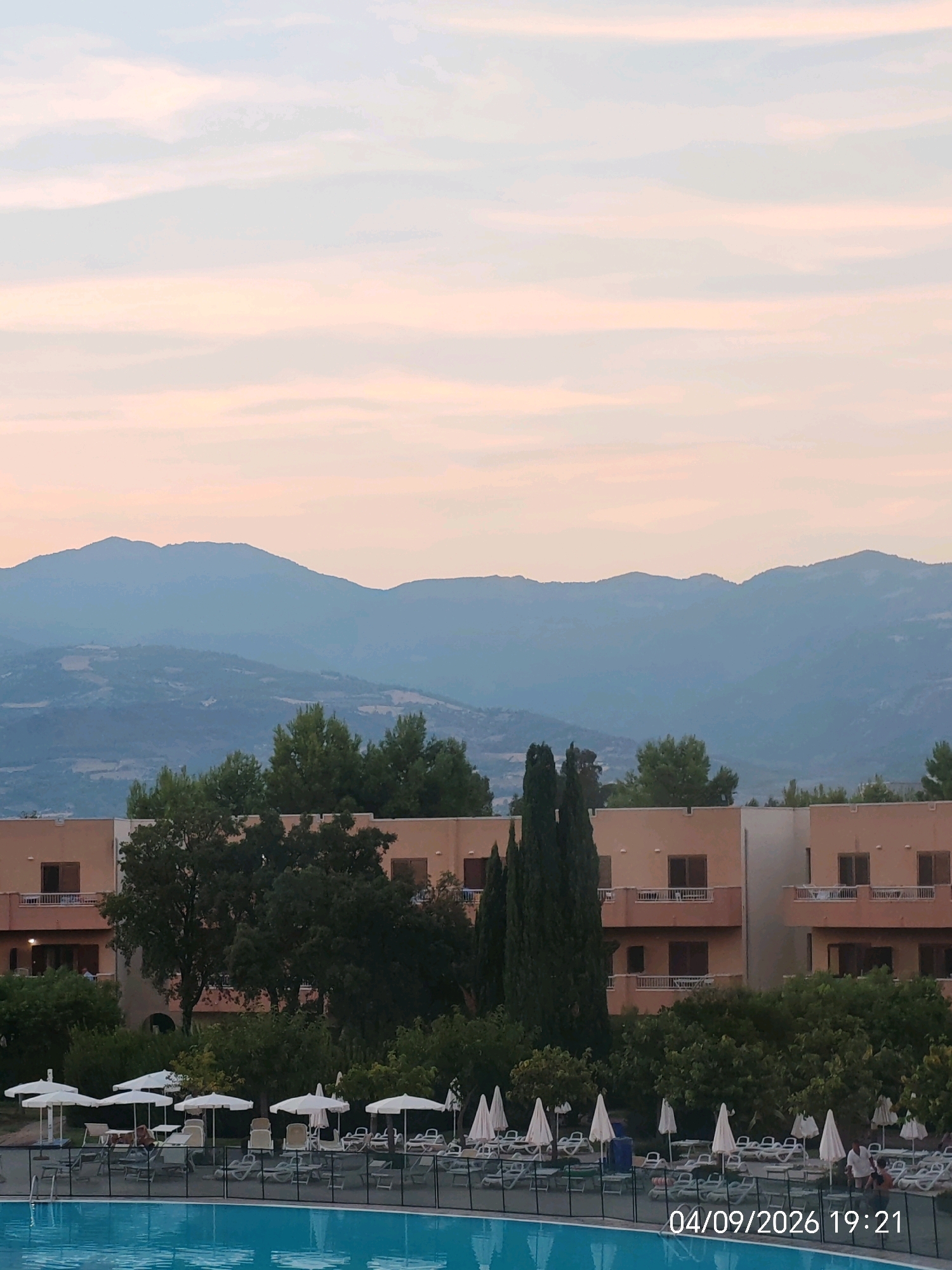 This image depicts a serene sunset view over a resort in Villapiana Scalo, Italy. The sky displays a gradient of soft pinks and pale blues, transitioning into twilight hues. In the foreground, a swimming pool is visible, surrounded by rows of empty lounge chairs and white umbrellas, suggesting a tranquil evening. Behind the pool area, a multi-story resort building stands, its windows casting a warm glow. Lush green trees and cypress trees flank the building, adding to the Mediterranean ambiance. In the distance, rolling hills and majestic mountains form the backdrop, their forms softened by the fading light. The timestamp "04/09/2026 19:21" is superimposed on the lower right portion of the image, indicating the date and time the photograph was taken. The overall scene evokes a sense of peacefulness and relaxation, characteristic of a holiday destination at dusk.