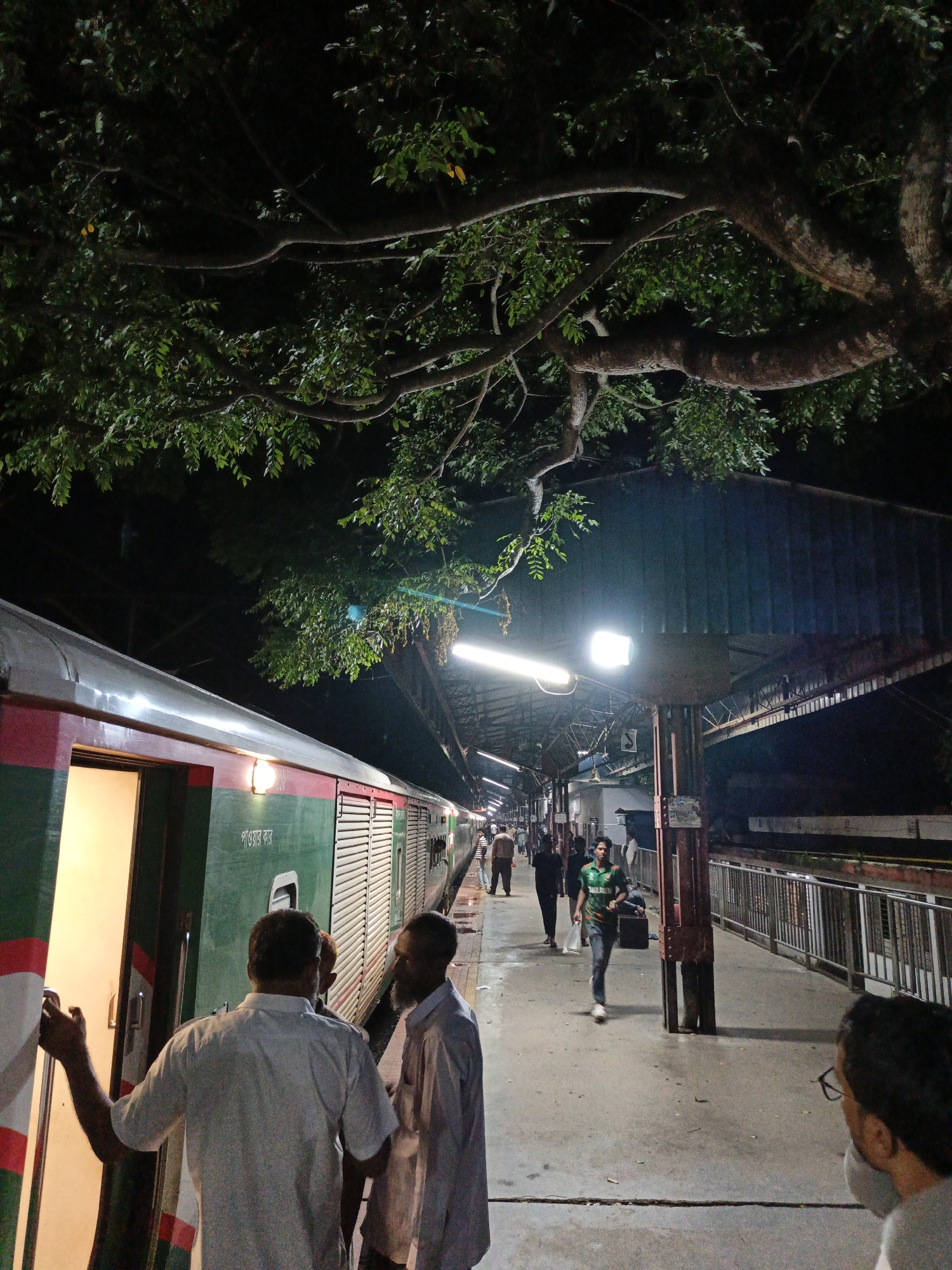 The image captures a scene at the Rajbari Railway Station in Rajbari, Bangladesh, at night. A train, predominantly green and red, is stopped at the platform with its door open, illuminated by warm interior lighting. Several individuals are present on the platform, with two men in the foreground appearing to be in conversation near the train door. Further down the platform, other passengers are walking, some carrying bags. Overhead, the branches of a large tree extend over the platform, with its leaves silhouetted against the night sky and the artificial lights of the station. The platform itself is paved and shows signs of recent rain, with some wet patches visible. The overall atmosphere is one of a busy transit point during evening hours.