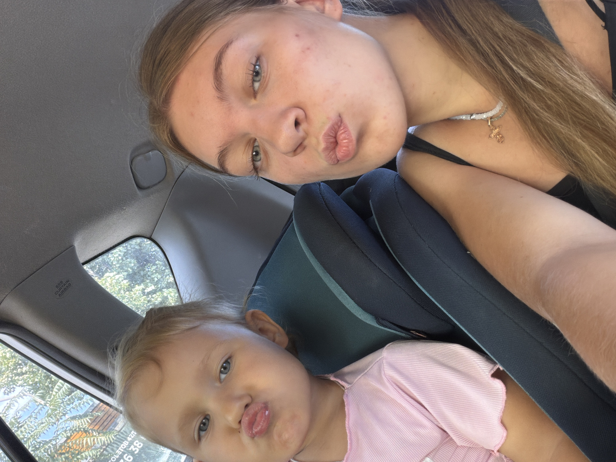 A close-up, selfie-style photo shows two young females in the back seat of a car. The person in the foreground, on the right, is an older female with blonde hair and blue eyes, wearing a black tank top and a necklace with a cross pendant. She is making a kissing face. To her left and slightly behind her is a younger female, likely a child, with blonde hair and blue eyes, wearing a pink shirt. She is also making a kissing face. The interior of the car is visible, including car seats and the headliner. The background outside the car window shows green foliage and a building. The photo is taken during the day.