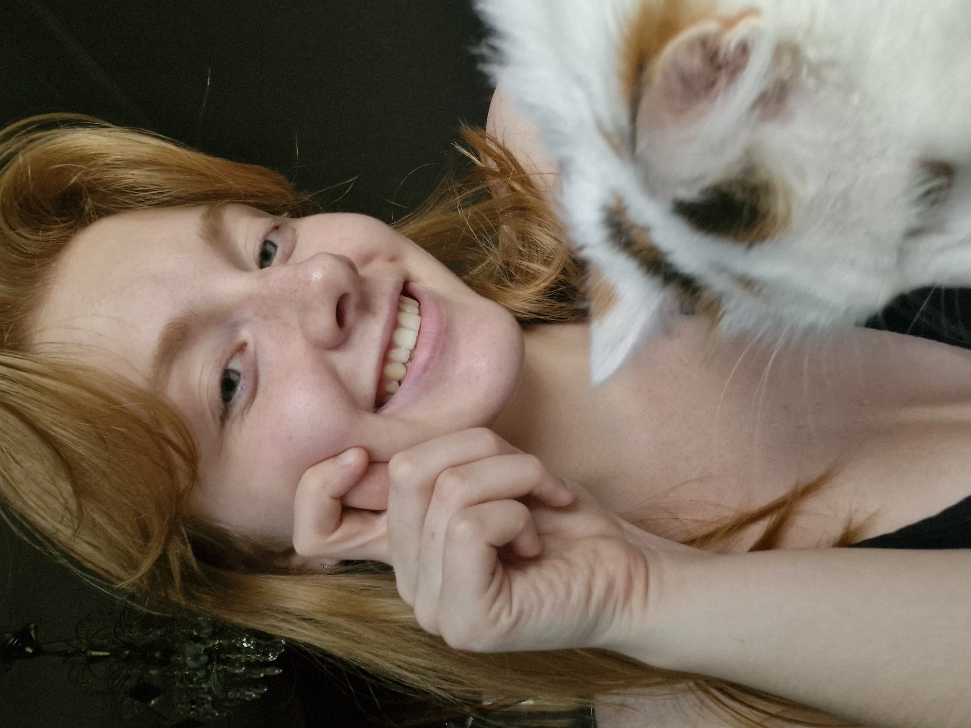 A young woman with red hair and freckles is posing for a selfie with a calico cat. She is smiling and holding her hand to her cheek. The cat's face is blurry and partially obscured by her hair. The background is dark and out of focus, with what appears to be a chandelier visible in the upper left corner. The image is a close-up shot, with the woman's face and the cat filling most of the frame. The setting is indoors, possibly a residential interior. There is no identifiable information about the location in Yekaterinburg, Russia.