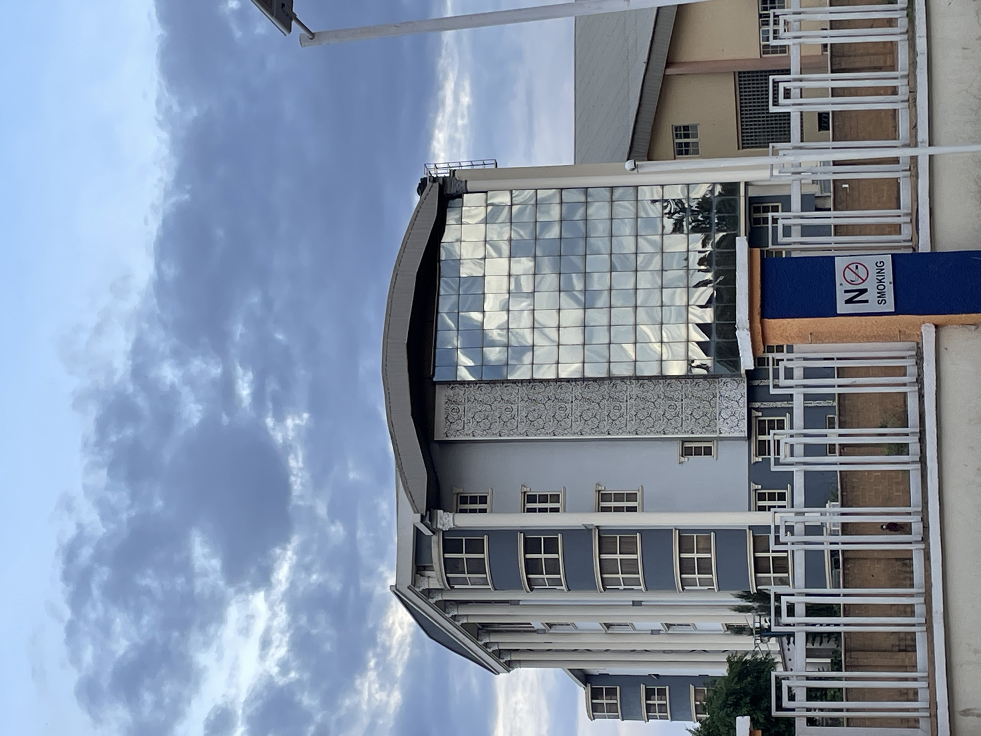 The image depicts a multi-story building in Jos, Nigeria. The building features a curved facade with a large glass block window. Lower floors have a blue-gray color with evenly spaced windows and white pillars. A white railing runs along the right side of the building, and a "No Smoking" sign is visible. The sky is overcast with blue sky peeking through, and a light pole is visible in the upper left.