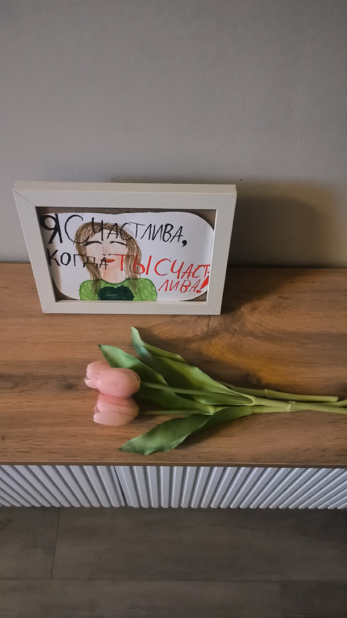 A framed drawing depicting a woman with brown hair and green clothing is displayed on a wooden shelf. The drawing has text written in Russian that translates to "I am happy when you are happy!". A bouquet of three pink tulips lies on the shelf in front of the frame. The background is a plain grey wall. The image appears to be taken indoors, possibly in a home environment. The location is not specified in the image, nor is it possible to determine the city of Kovrov, Russia from visual cues.