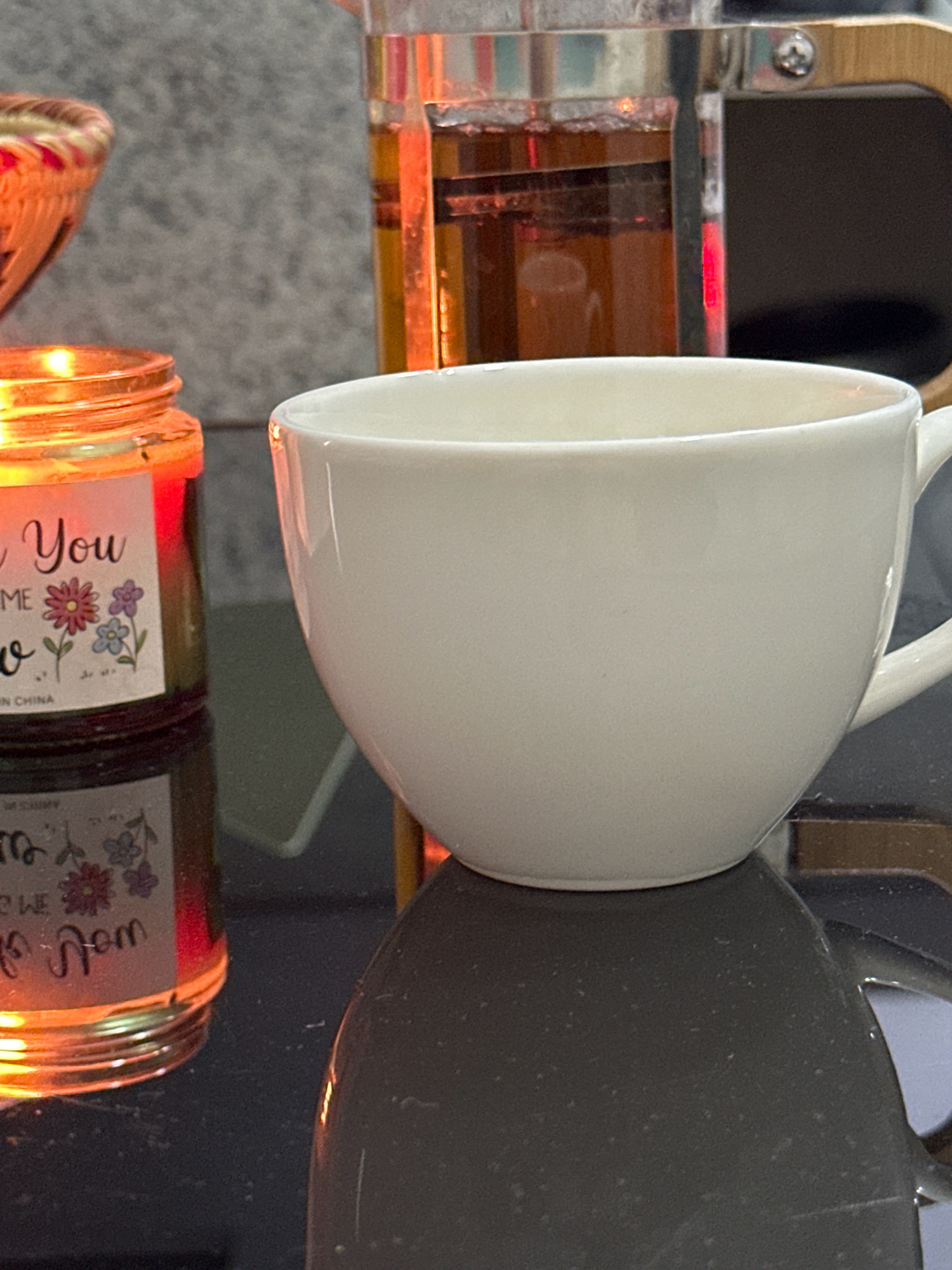 In Lalitpur, Nepal, a close-up shot captures a serene moment. A white ceramic teacup rests on a dark, reflective surface, its smooth curves catching the warm light. To the left, a lit candle in a glass jar emanates a soft orange glow, illuminating a label with floral motifs and the partially visible text "You". Behind the cup and candle, a glass French press filled with amber-colored liquid suggests brewed tea. The background is a mottled gray texture, adding to the tranquil ambiance. The scene is devoid of any human presence, focusing solely on the objects and their interplay of light and shadow, evoking a sense of quiet contemplation.