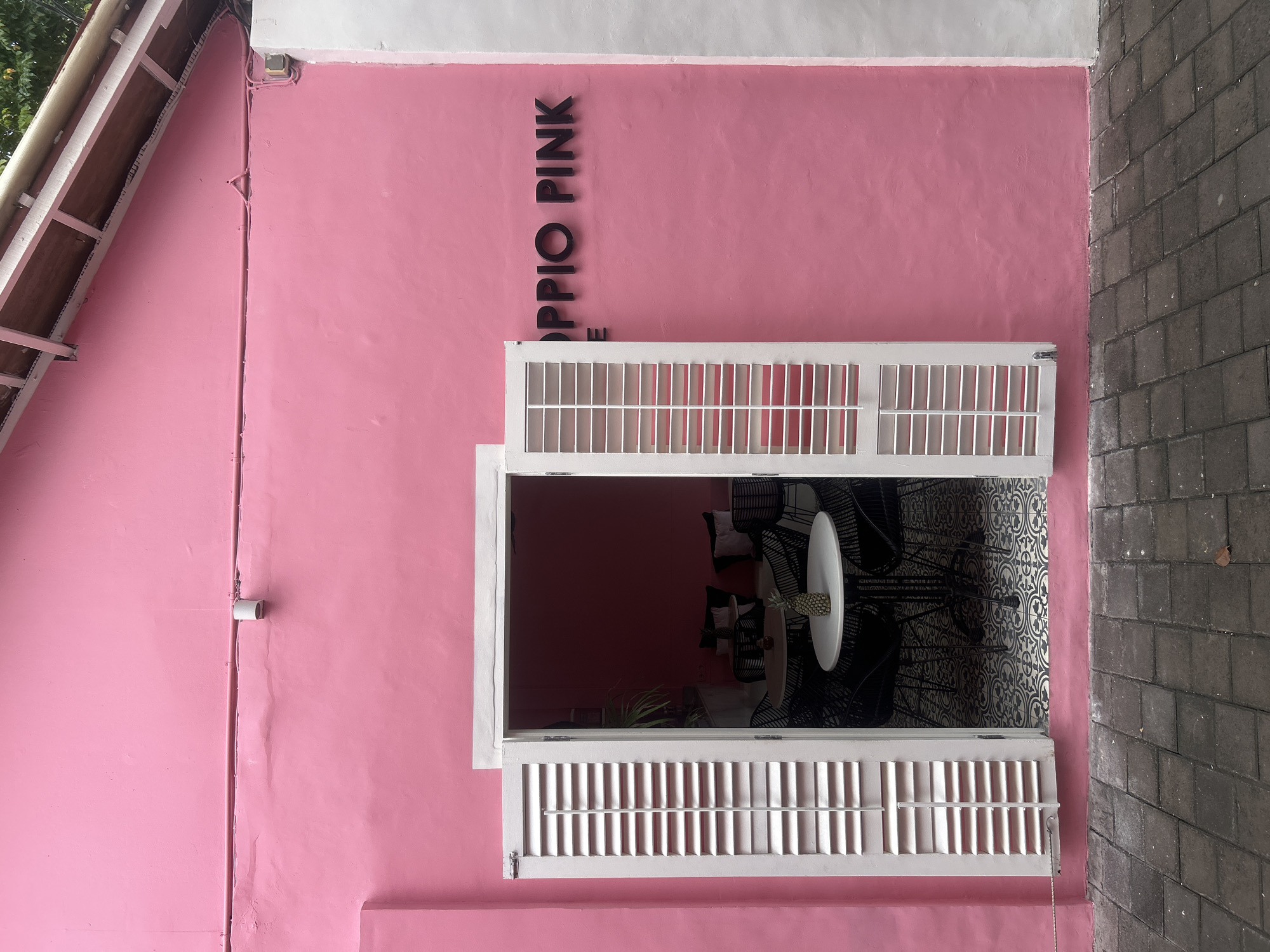 The exterior of a brightly painted pink building is shown, with a white-framed doorway opening into a cafe interior. The doorway features white louvered shutters, with the left shutter ajar. Inside, round tables with black chairs are visible, some adorned with black cushions. A small pineapple sits on a table near the entrance. Black lettering on the pink exterior wall reads "OPPIO PINK". The building is situated on a cobblestone pathway. The scene appears to be in Kuta, Indonesia, given the architectural style and lush greenery visible in the background.