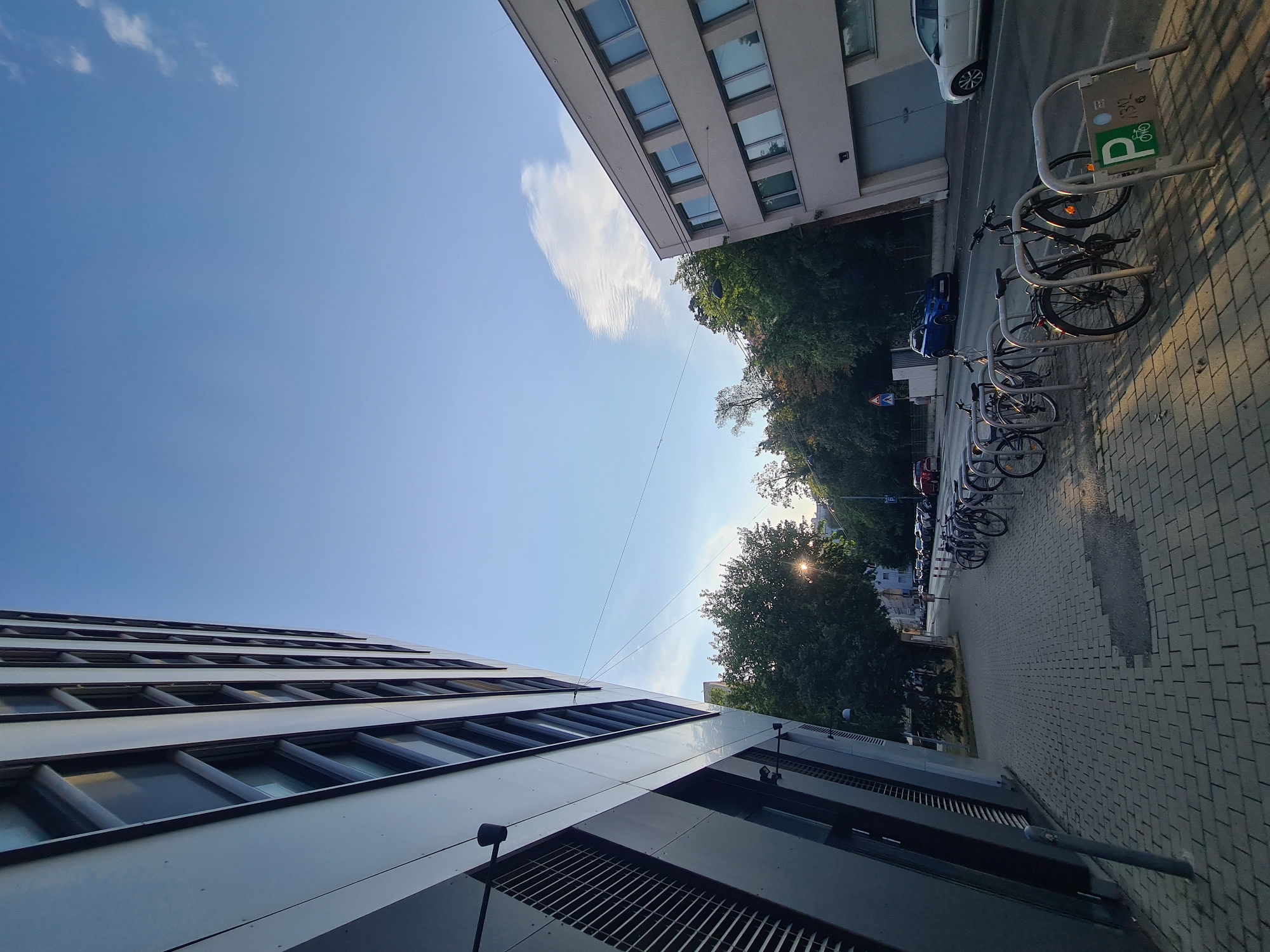 This is a street-level, wide-angle photograph taken in Vienna, Austria, looking down a sidewalk lined with trees and buildings. On the left, a modern, multi-story building with a dark gray facade and rows of windows dominates the foreground. The building is angled, giving a dynamic perspective to the shot. The sidewalk in front of it is paved with light gray rectangular bricks. To the right of the sidewalk, a row of bicycles is parked in a metal rack. Beyond the parked bicycles, a street runs parallel to the sidewalk, with cars parked along the side and a few vehicles driving. One car is a blue SUV, and another is a white sedan. Further down the street, more trees and buildings are visible. The sky above is a clear, pale blue with a few wispy white clouds. The sun is low in the sky, casting long shadows and creating a warm, golden light that illuminates the trees and the bicycles. A pedestrian crossing signal is visible in the distance. The overall impression is a quiet urban scene, likely in the late afternoon or early evening.