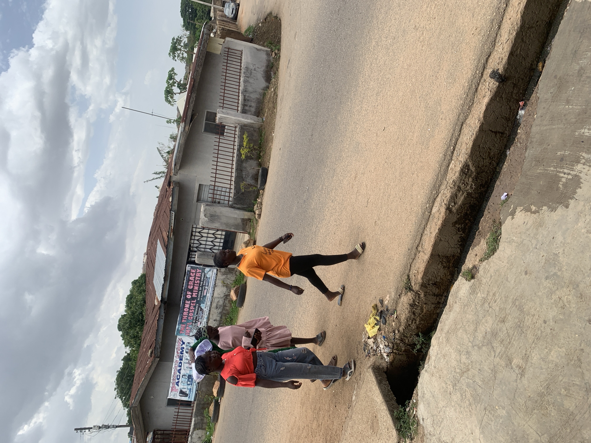 The image captures an outdoor scene in Jos, Nigeria, depicting three individuals walking along the side of a paved road next to a concrete drainage ditch.
The person closest to the camera is wearing a red top, jeans, and sneakers. She is followed by another person wearing a light pink dress and a head covering. The third person wears an orange T-shirt and dark pants.
Buildings with corrugated metal roofs line one side of the road. Signage for "PRECIOUS STONE ACADEMY" and "THE THRONE OF GRACE INTL GOSPEL MINISTRY" are visible on the buildings. The sky is overcast with white and gray clouds. A few trees are seen in the background. The road is well-worn. There is some litter and debris in the drainage ditch.