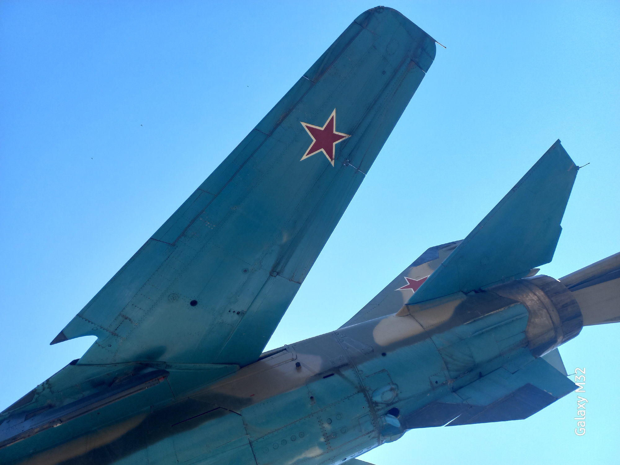 The image shows the tail section of a military jet, possibly a MiG-21 or similar Soviet-era aircraft, painted in a blue and brown camouflage scheme. The tail fins are prominently visible, each bearing a red star, a symbol of the former Soviet Union. The aircraft appears to be on display, possibly as a monument. Given the visual cues and the aircraft's history, a location in Sochi, Russia is probable, though precise confirmation would require additional information. The sky is clear blue. The scene is taken from a low-angle perspective looking up at the aircraft.