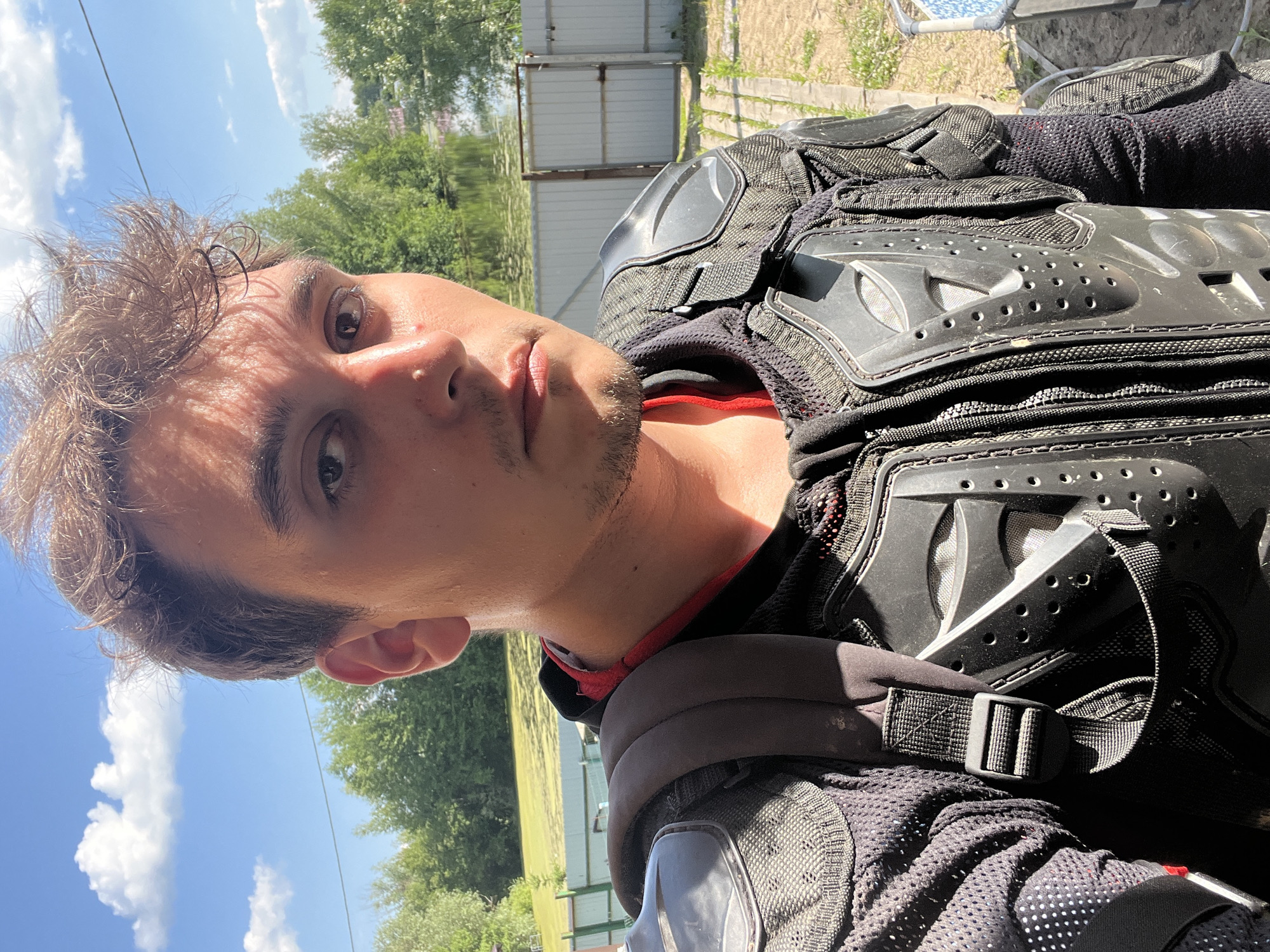 A selfie taken outdoors in Vasilyevo, Russia, features a young man wearing a black padded motocross jacket. He is positioned in the foreground, facing the camera directly, with a neutral expression. The background shows a body of water, lush green trees, a light-colored fence, and a bright blue sky with scattered clouds. The lighting suggests a sunny day.