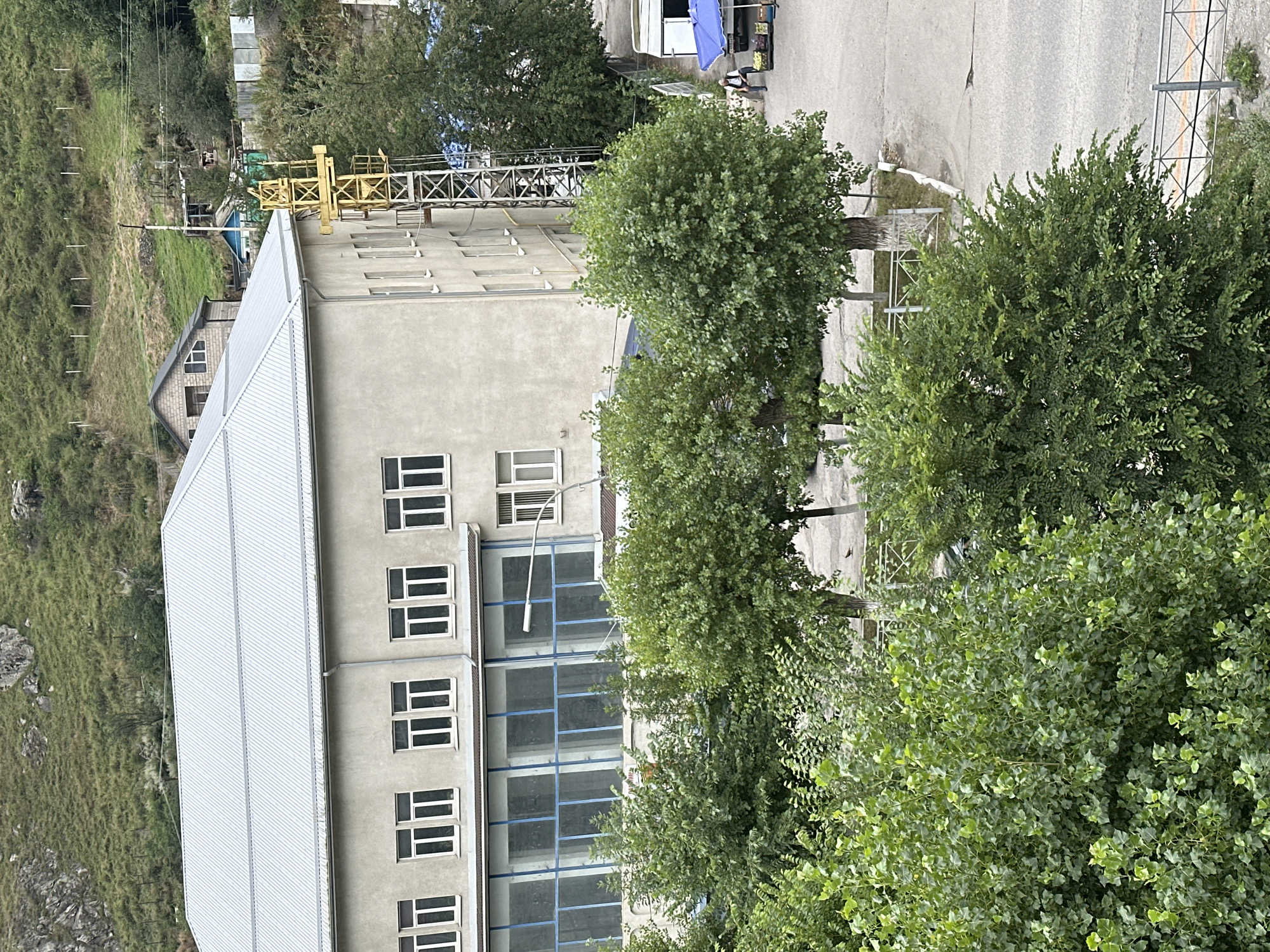 The image, taken in Tyrnyauz, Russia, depicts a multi-story building with a light-colored, stucco exterior and a corrugated metal roof. The building features numerous rectangular windows on its upper floors and a long, glass-enclosed section on the ground level with blue-tinted window panes. A yellow metal structure, possibly a crane or scaffolding, is attached to the side of the building. In the foreground, lush green trees and bushes obscure the lower portion of the scene. Beyond the building, a street is visible with a white trailer and a blue umbrella shading a small market stall where a person is standing. The background shows a steep, grassy hillside with scattered trees and some distant structures. The overall environment appears to be a somewhat rural or semi-urban setting, possibly during daylight hours under an overcast sky.