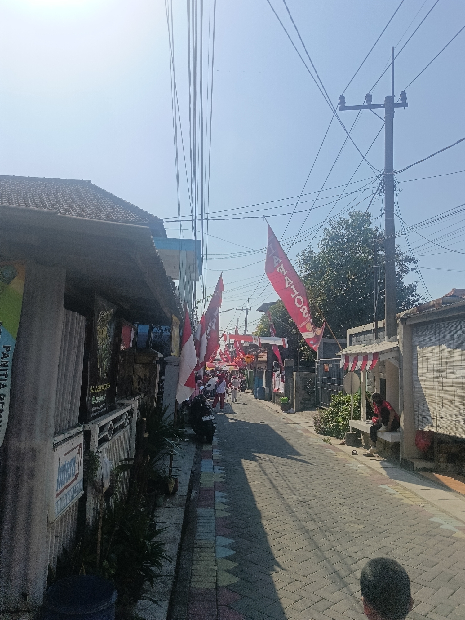 The image depicts a street scene in Gedangan, Indonesia, under a clear sky. The street is paved with interlocking bricks and lined with houses. Several Indonesian flags, red and white, are displayed prominently along the street, indicating a possible celebration or event. One large red flag with white text, partially obscured, hangs from a pole on the right side of the street. In the foreground on the left, a building with a dark-tiled roof is visible, featuring a banner with green and white text. Further down the street, a group of people, some in white clothing, appear to be walking or participating in an event. A motorcycle is parked on the side of the road. A power pole with multiple wires stretches across the upper portion of the image. On the right side of the street, a person is sitting on a porch. The overall impression is of a residential street decorated for a special occasion.