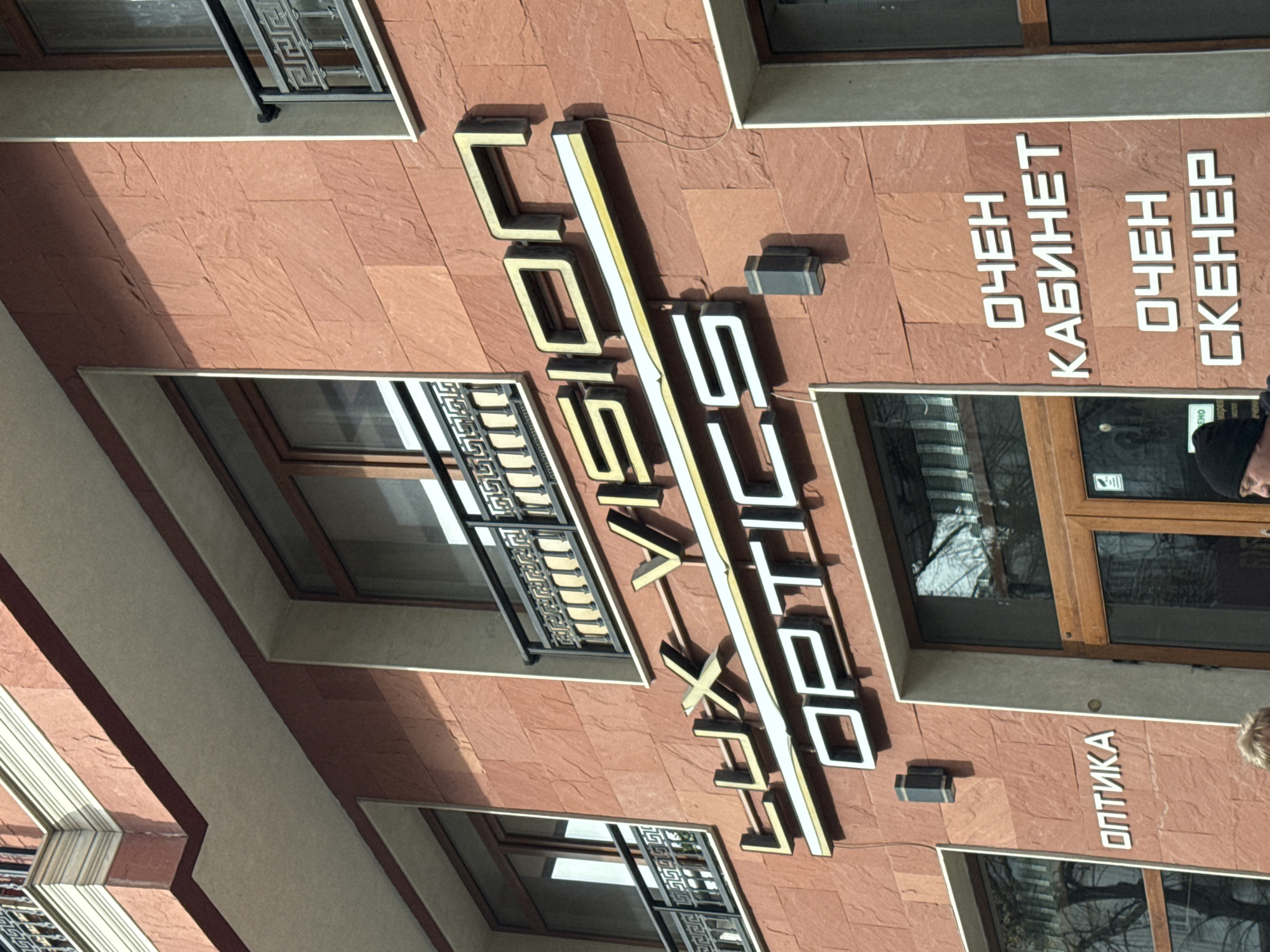 The image captures the exterior of an optometry clinic in Varna, Bulgaria. The building is constructed with textured, reddish-brown panels. The signage, rendered in a modern, stylized font, displays "COLVIS LUX OPTICS" vertically. To the right of the logo, text in Cyrillic script indicates "OPTICS," "EYE EXAM ROOM" (transliterated), and "EYE SCANNER" (transliterated). The building has a window with a black decorative security grill. The window is partially visible due to a person standing to the right of the building. The photo appears to be taken during daylight hours.