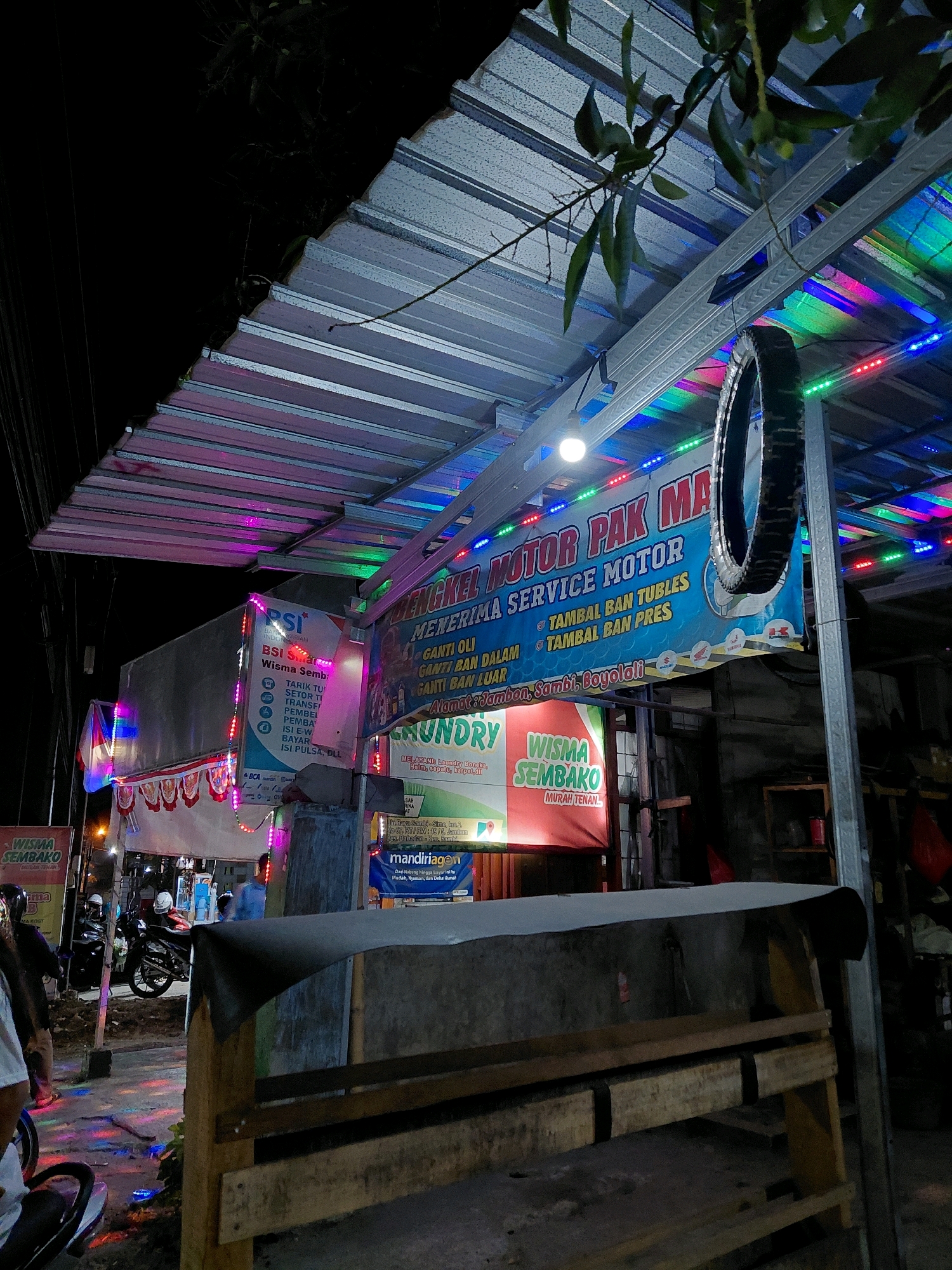 The image depicts a night scene on a street in Babadan, Indonesia. The foreground shows a wooden structure and a street, with some colorful lights reflecting on the ground. In the midground, there are several signs and businesses illuminated by string lights, including "WISMA SEMBAKO," "LAUNDRY," and a banner advertising "ENERIMA MOTOR PAK MBR" with services like "GANTI OLI" and "TAMBAL BAN." The BSI logo is also visible on a building. Further back, there are people and motorcycles, suggesting a busy commercial area. The environment is characterized by artificial lighting, typical of a nighttime street in a town.