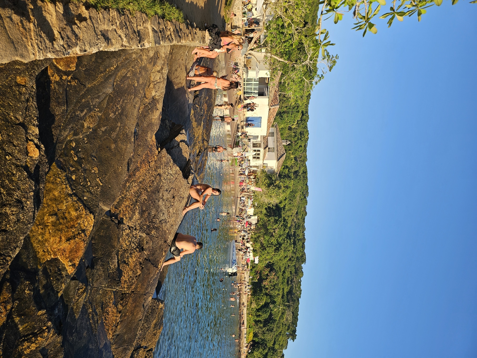 The image depicts a beach scene in Armação Dos Búzios, Brazil. A rocky cliff occupies the left side, with two people climbing on it. A woman in a bikini sits near the edge, and a man in dark shorts is positioned lower down.
The beach is crowded with people swimming, sunbathing, and walking along the shore. Buildings, including white structures with red tile roofs, are visible behind the beach. The shoreline is lined with lush green vegetation. The sky is a clear, bright blue. People are observed sunbathing on the beach and swimming in the water. The sun appears to be setting, casting long shadows on the rocks.