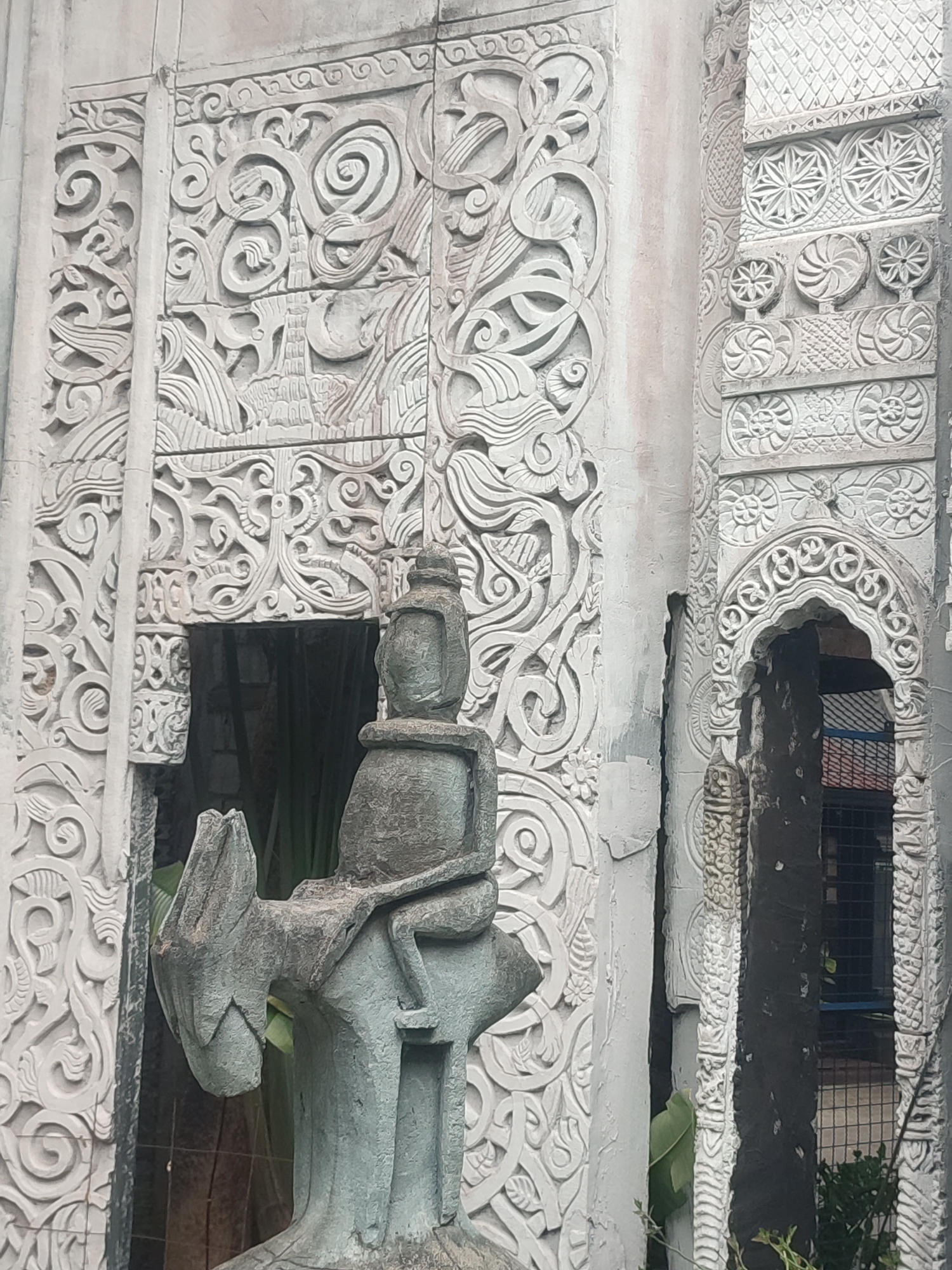 The image shows a building with intricate, ornate carvings. A gray statue of a figure riding an animal, possibly a donkey or stylized quadruped, stands in front of the building. The statue is weathered. The building's exterior is off-white or light gray, with detailed carvings that appear to be floral or abstract designs. There are arched entryways on the right side of the image, and some greenery is visible at the bottom. The scene is located in Muang Pattaya, Thailand, as specified in the prompt. The style of carving suggests a temple or religious structure.