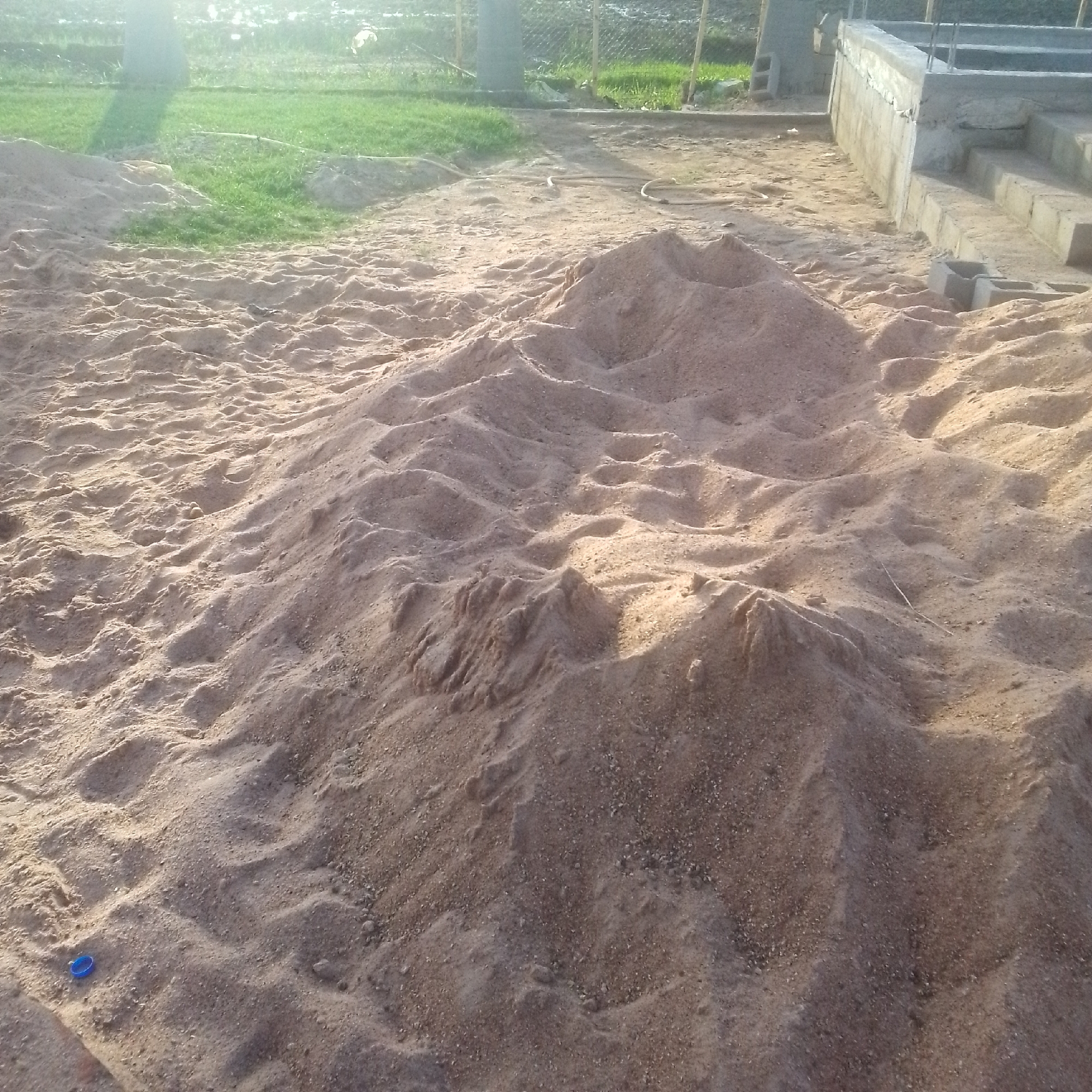 The image depicts a construction site in Jalingo, Nigeria. The primary subject is a large pile of sand in the foreground, with distinct undulating textures and foot impressions visible. In the background, to the right, a concrete structure is under construction, featuring steps and reinforcement bars. Adjacent to the sand pile, a small blue object, possibly a cap or a piece of plastic, is noticeable. Further in the background, a grassy area and a chain-link fence are visible, suggesting an outdoor, possibly semi-rural or developing urban environment. The lighting suggests it is daytime, with a bright, possibly sunny, atmosphere that casts shadows. The overall scene conveys a sense of ongoing construction work, with materials prepared for building. No people are visible in the frame, and no text is discernible.