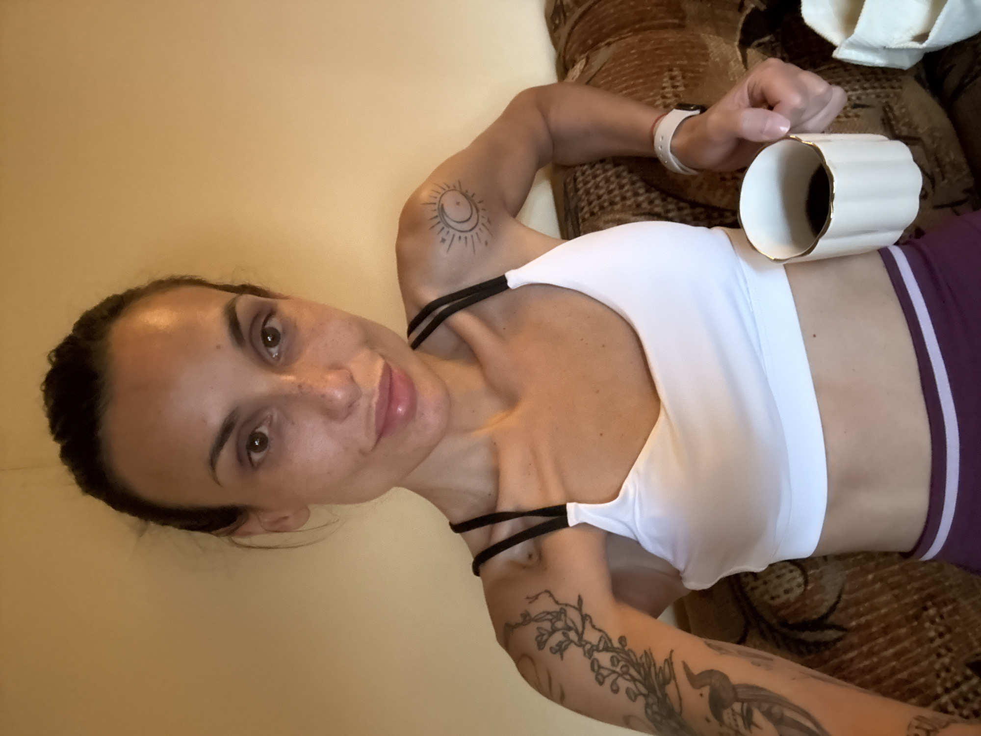 A woman is captured in a close-up, high-angle shot, lying down indoors, likely on a couch or bed. She looks directly at the camera with a relaxed, gentle smile, her brown eyes engaged. Her dark hair is pulled back, and she has visible freckles across her nose and cheeks. She is wearing a white sports bra with black straps and purple bottoms featuring a white stripe.
Numerous tattoos adorn her arms; a sun and moon design is visible on her left bicep, and a botanical pattern extends down her left forearm. On her right wrist, she wears a white wristband. She holds a white, fluted mug with a gold rim in her right hand, seemingly taking a moment of repose with a drink.
The background consists of a plain light brown wall on the left and a textured, patterned brown upholstered surface on the right. The overall lighting is soft, suggesting an indoor daytime setting. No visible text is present in the image. The scene takes place in Pleven, Bulgaria.