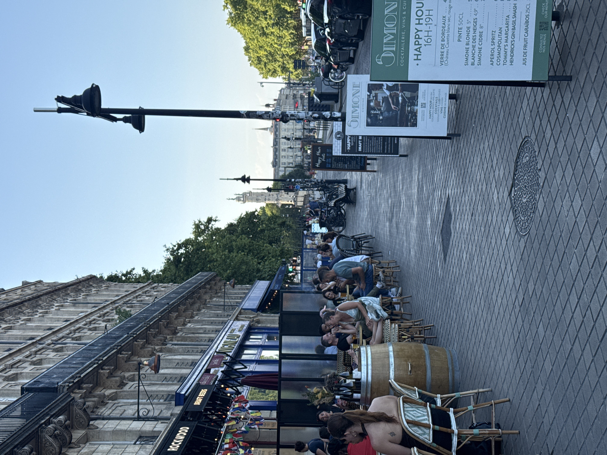 This image depicts a lively outdoor dining scene in Bordeaux, France, likely on the Quai de Bacalan or a similar waterfront promenade given the architecture and proximity to the Garonne river. The time appears to be late afternoon or early evening, judging by the light and the presence of happy hour signs.
In the foreground, several patrons are seated at tables and chairs set up on a patterned stone sidewalk. They are engaged in conversations and enjoying drinks. A large wooden barrel is positioned near some of the tables, adding to the rustic ambiance. A woman with a tattoo on her back is seated at a table in the lower left of the frame.
Along the sidewalk, a series of A-frame signs advertise "Happy Hour" specials and menu items from the establishments lining the street. The signs prominently feature the name "SIMONE" along with details of cocktails, wines, and other offerings. One sign shows a culinary scene with a chef preparing food.
The background reveals a grand, Haussmannian-style building with multiple stories, ornate balconies, and arched windows, characteristic of Bordeaux's architecture. Trees line the street, providing shade and greenery. In the distance, city landmarks such as a tower and other buildings are visible, suggesting a central urban location. Several cars are parked along the street, and a bicycle rack with parked bicycles is also present. The sky is clear and blue.