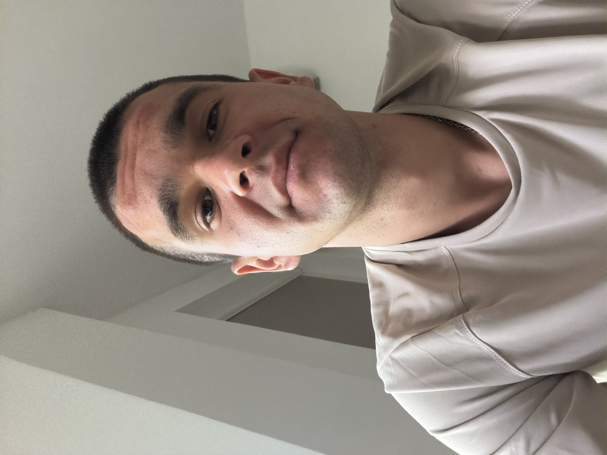 An upside-down portrait features a young adult male looking directly at the camera with a subtle smirk or wry expression. He has short, dark hair, dark eyes, and fair skin. He is wearing a light beige or tan long-sleeved shirt with visible stitching details around the shoulders and collar. A thin, silver-toned chain is visible around his neck, resting just below the collar of his shirt.
The image is captured from a low angle, making the subject appear to be looking upwards into the lens, suggesting a selfie or a shot taken from below. A faint reddish mark is visible on his forehead. The background is simple and uncluttered, consisting of plain, light-colored (white or light grey) walls and a ceiling, indicative of an indoor setting, possibly a room in a residence in Veliko Tarnovo, Bulgaria. Architectural elements, such as a door frame or wall corner, are subtly visible in the background. The lighting is soft and even, illuminating the subject's face without harsh shadows. There is no visible text in the image.