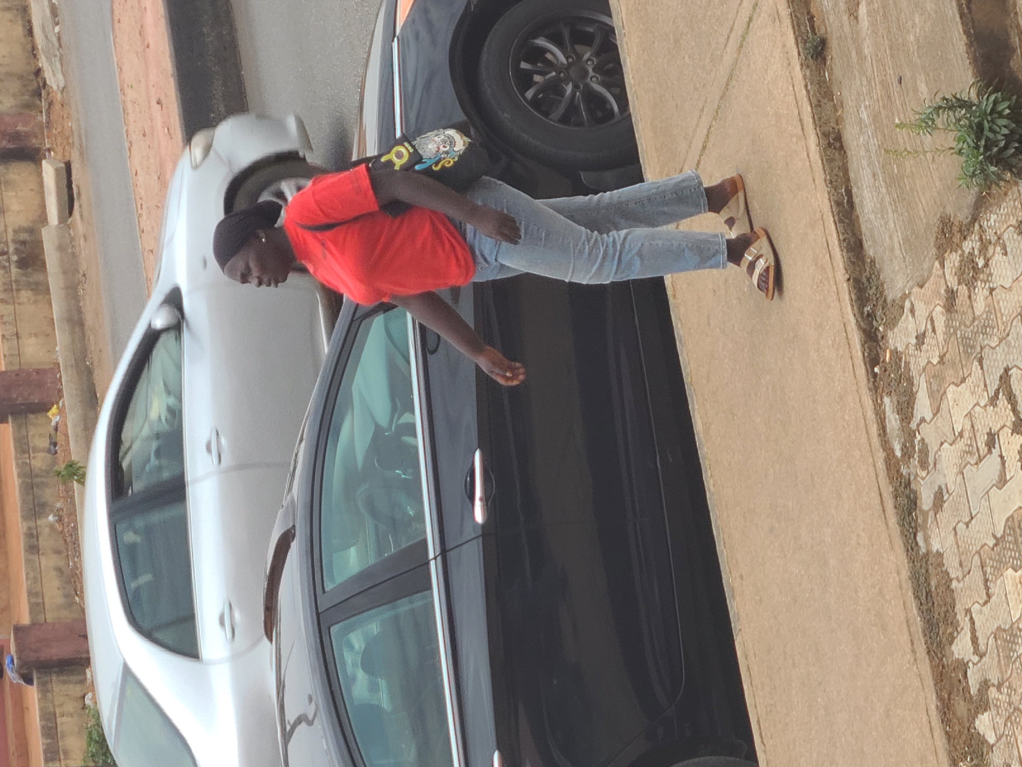 A woman stands on a sidewalk next to two cars. The woman is wearing a red t-shirt, blue jeans, and sandals. She is holding a handbag. The cars are parked on a street. The sidewalk is made of concrete, and the street is paved. The location is in the city of Jos, Nigeria.