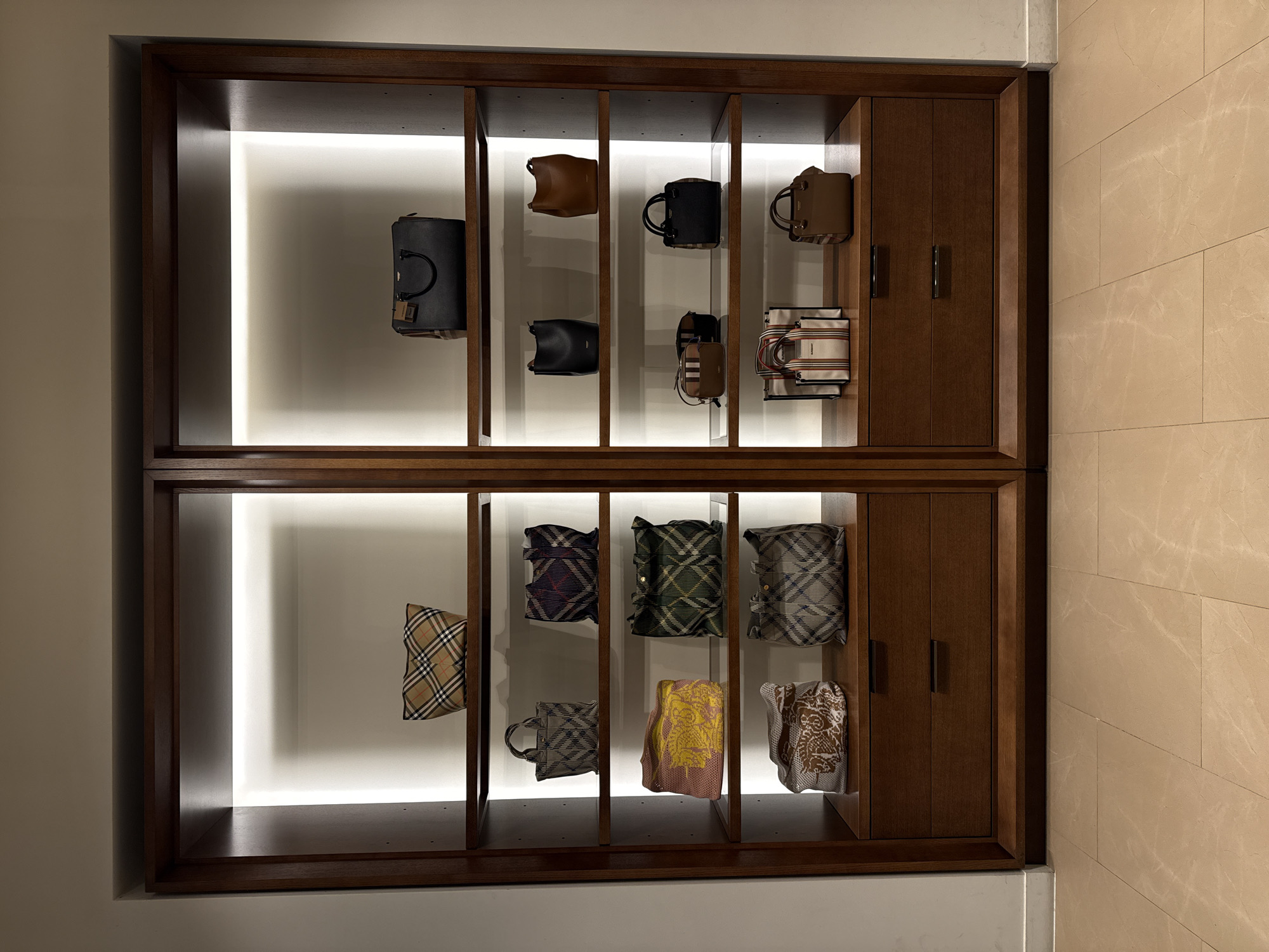 The image displays a wooden display case filled with handbags. The case is divided into compartments by vertical wooden supports. Some compartments have a bright light behind them, highlighting the handbags. The handbags vary in style, color, and material. Some appear to be structured handbags, while others are softer and more casual. The display case seems to be mounted on a wall. The overall aesthetic is clean and modern. The location in Gimpo-si, South Korea is not determinable from the image.