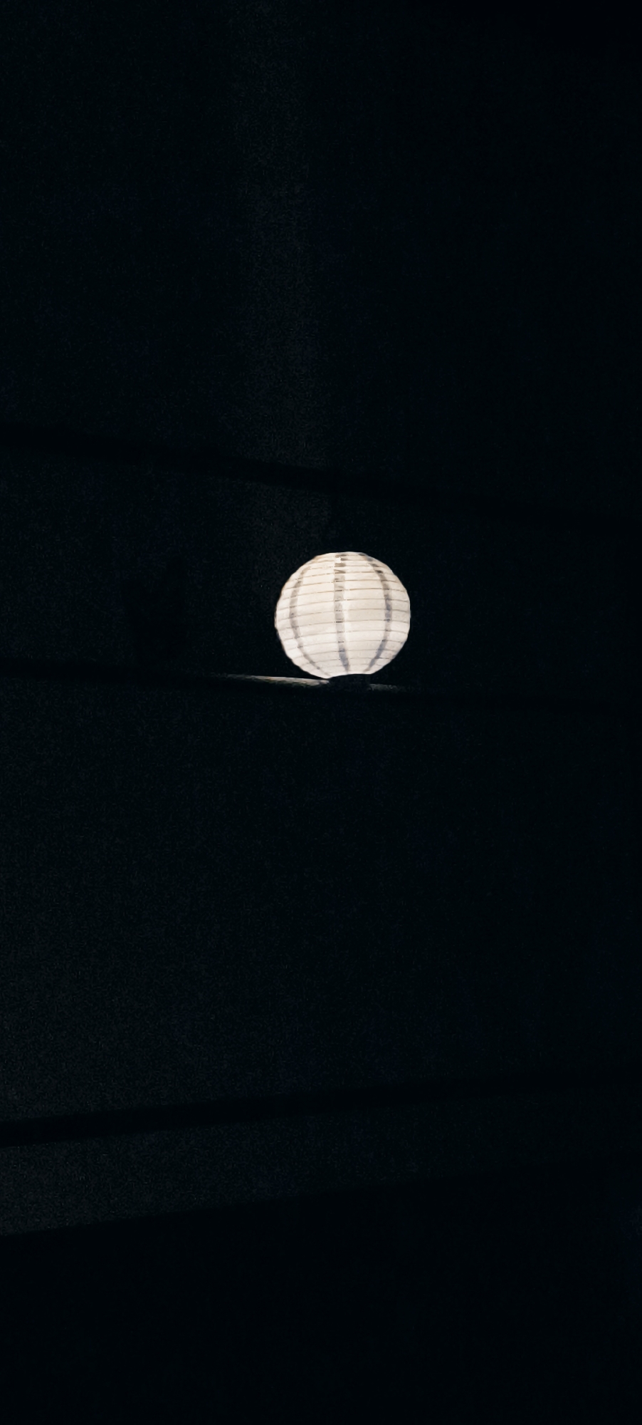 A white, spherical paper lantern with horizontal stripes of darker material is illuminated from within, casting a soft glow against a dark, textured background. The lantern is positioned centrally in the frame, with subtle shadows extending outwards. The environment appears to be indoors or in a dimly lit outdoor setting, possibly a dimly lit area in Winnenden, Germany. There are no people or other discernible objects in the image.