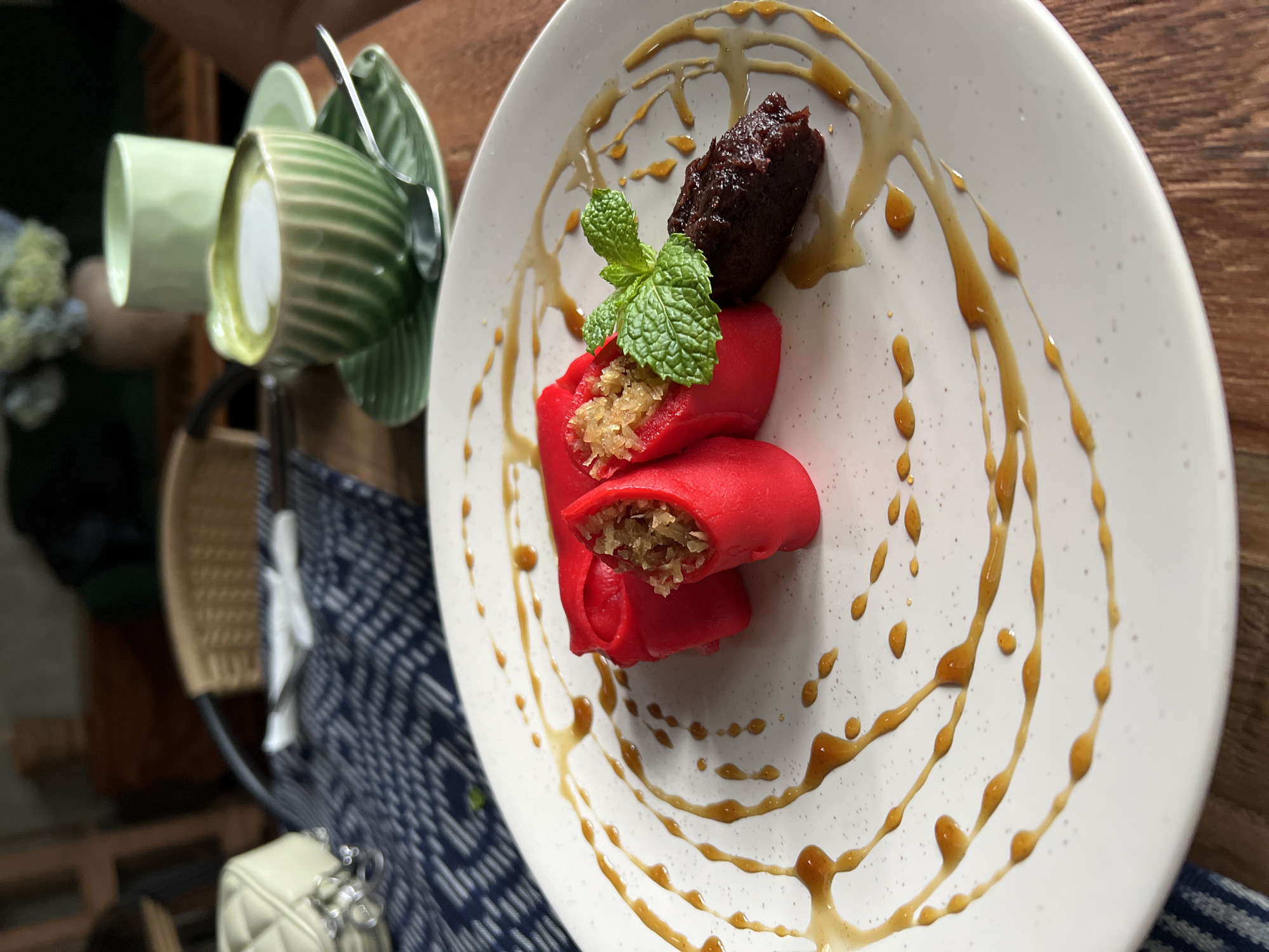 The image shows a dessert plate in a restaurant setting, likely in Singakerta, Indonesia. The plate contains several red-colored rolls filled with shredded coconut, garnished with a mint sprig, and accompanied by a dark brown paste or spread. The plate is decorated with concentric circles of caramel sauce. Beside the plate, there are green teacups stacked on top of each other with a spoon laying on top. In the background, there is a wooden table and a blue and white patterned cloth, and a white purse. The image appears to be taken in a well-lit, casual dining setting.