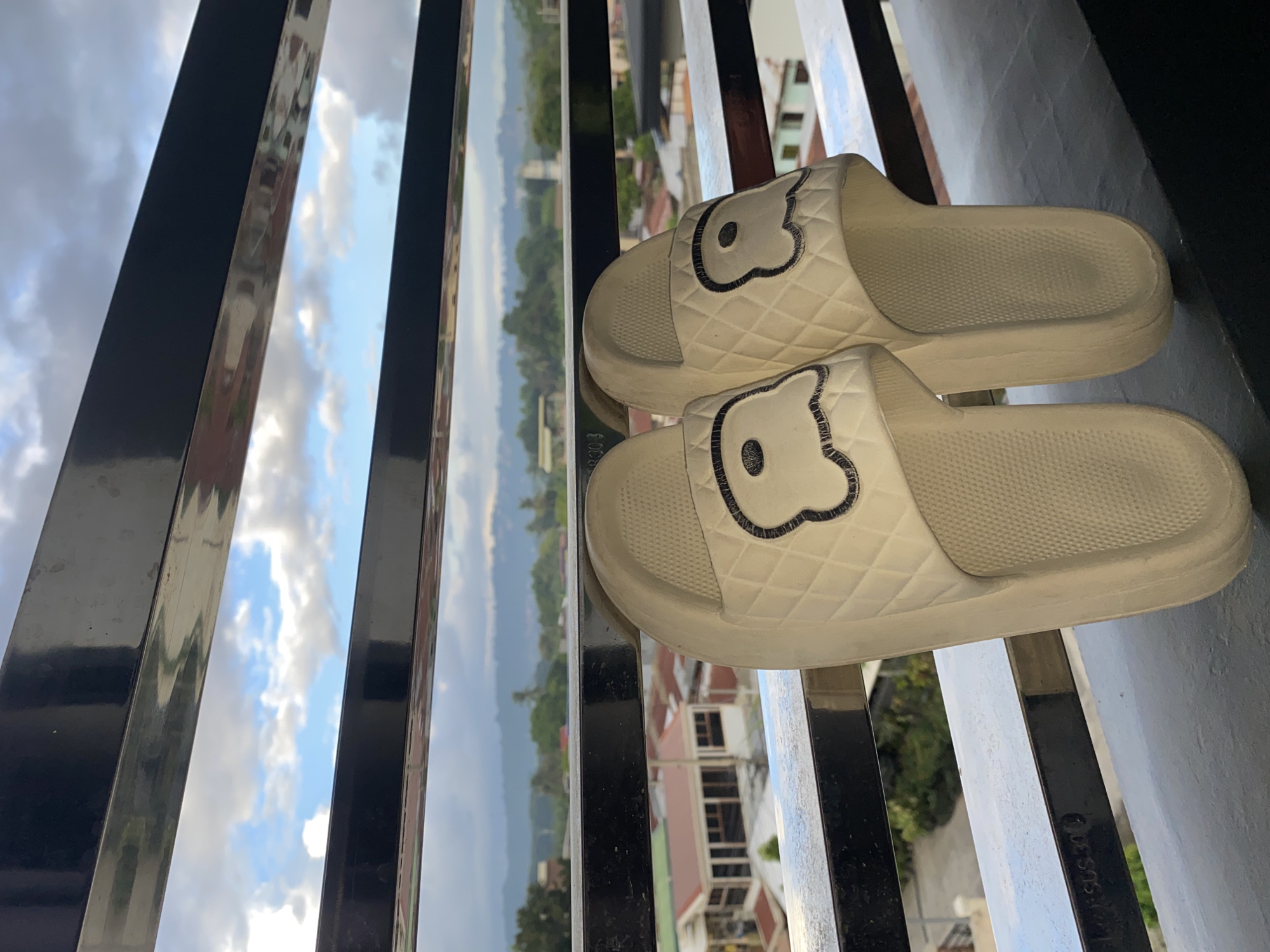 Two white platform slides with a quilted pattern and a black outline of a teddy bear face are positioned on a balcony railing. The slides are white with a textured footbed and a thick sole. The background shows a cityscape with buildings, trees, and a cloudy sky, indicative of Davao City, Philippines. The scene is captured from a slightly elevated perspective, looking out over the city.