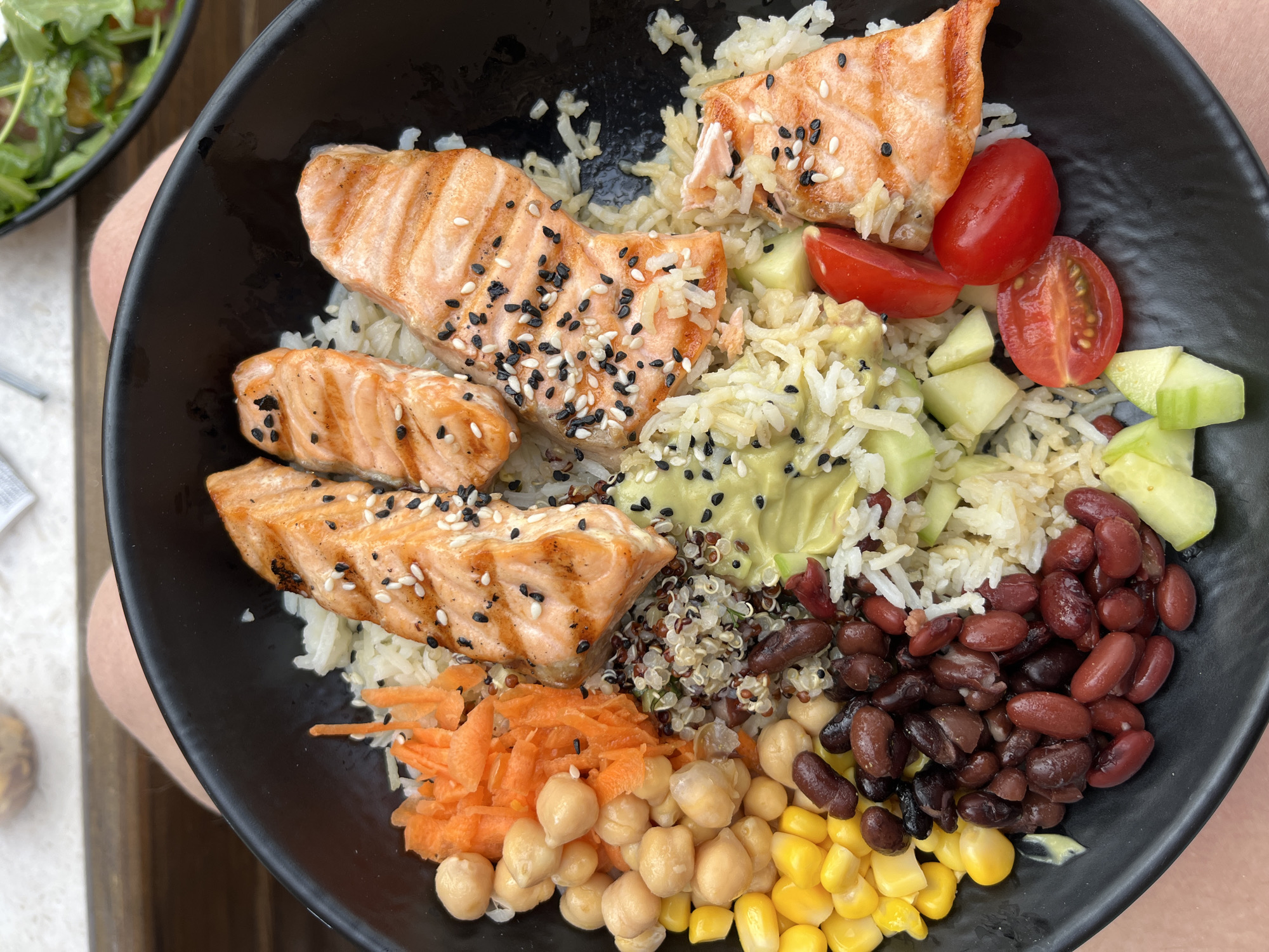 A close-up, overhead shot shows a black bowl filled with a salmon poke bowl. The bowl contains white rice, grilled salmon pieces seasoned with black and white sesame seeds, chickpeas, shredded carrots, corn, kidney beans, quinoa, diced cucumber, mashed avocado, and cherry tomato halves. The bowl is placed on a person's lap, suggesting an outdoor seating arrangement, possibly at a restaurant in Thassos, Greece, given the context. The lighting is natural and bright, highlighting the textures and colors of the food. The background is blurred but hints at a wooden surface and a glimpse of green foliage.