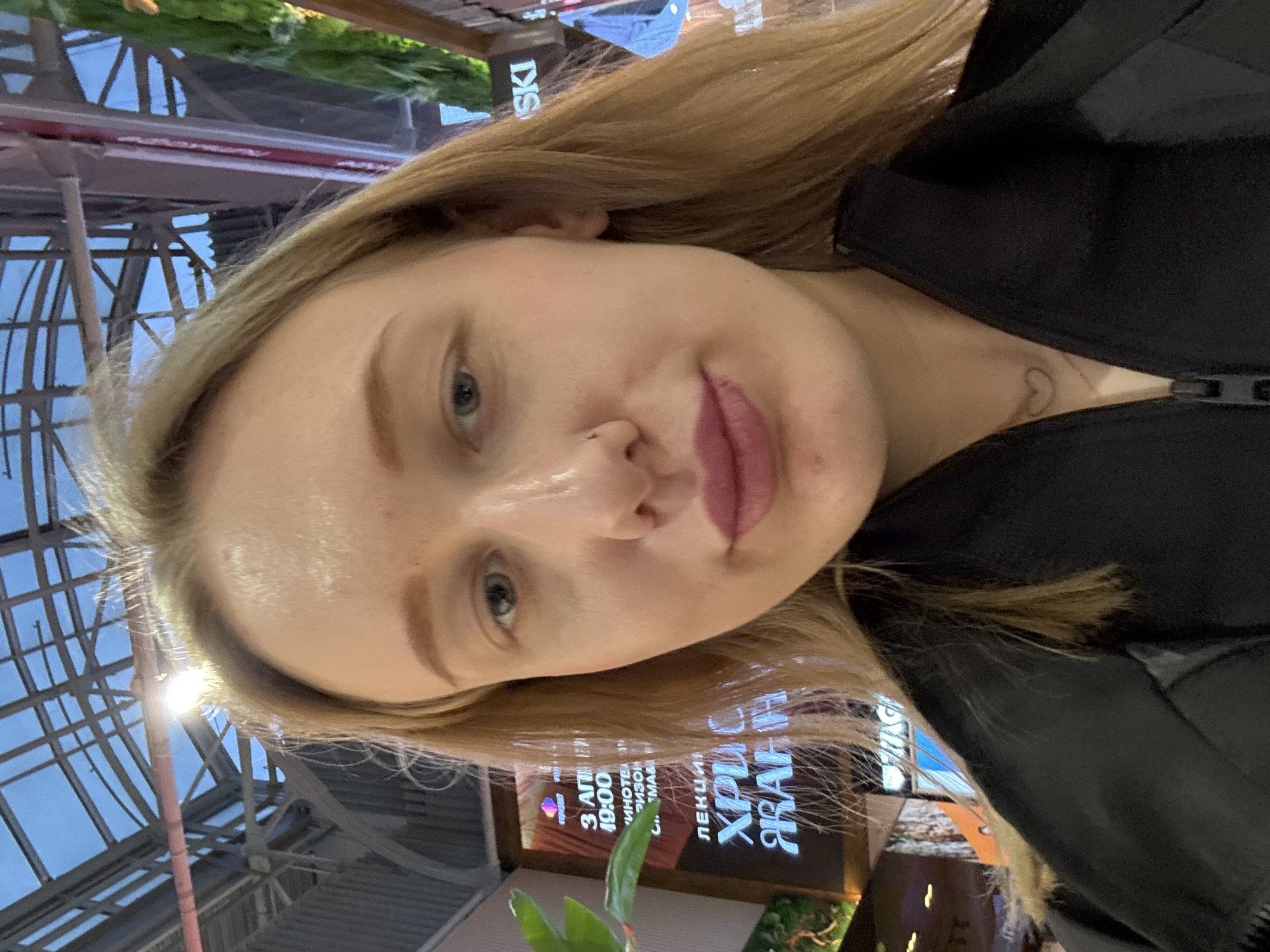 A selfie of a blonde woman with a nose piercing is the primary subject. She is wearing a black jacket, and a small heart tattoo is visible on her neck. The background shows a glass-roofed structure and signs with text, including "ЛЕКЦИ ХРИС ЖАНН," "3 ΑΠΗ 19:00," and "SKI." The context suggests an indoor location, possibly in Ol'ginskaya, Russia, given the Cyrillic text on the signs.