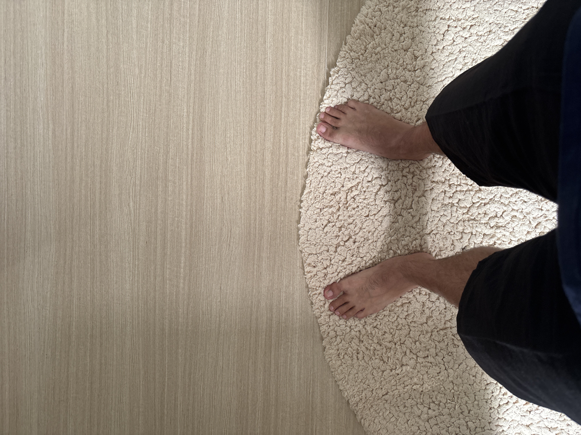 An overhead, eye-level shot shows a person's bare feet standing on a cream-colored, fuzzy rug that is curved along the bottom edge of the frame. The rug sits on a light-colored wooden floor. The person is wearing black shorts. The top portion of the image is dominated by the wood grain of the floor, with the rug occupying the bottom half.